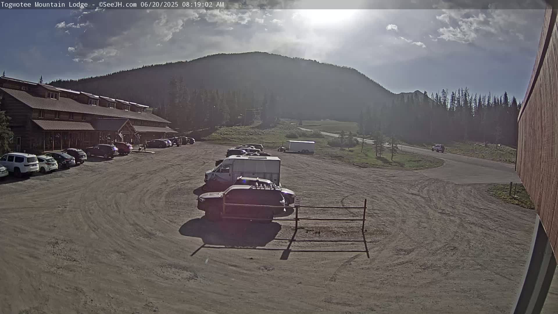 A long, low building with a parking lot full of cars sits in front of a large mountain under a partly sunny sky.