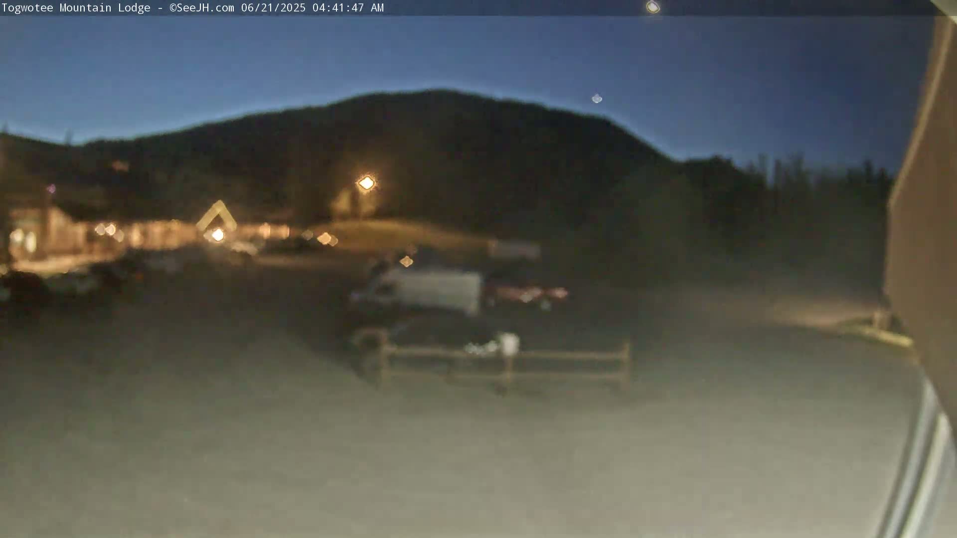 A dark, blurry nighttime scene shows a parking lot with vehicles in front of a lodge and a hill in the background under a clear, dark sky.