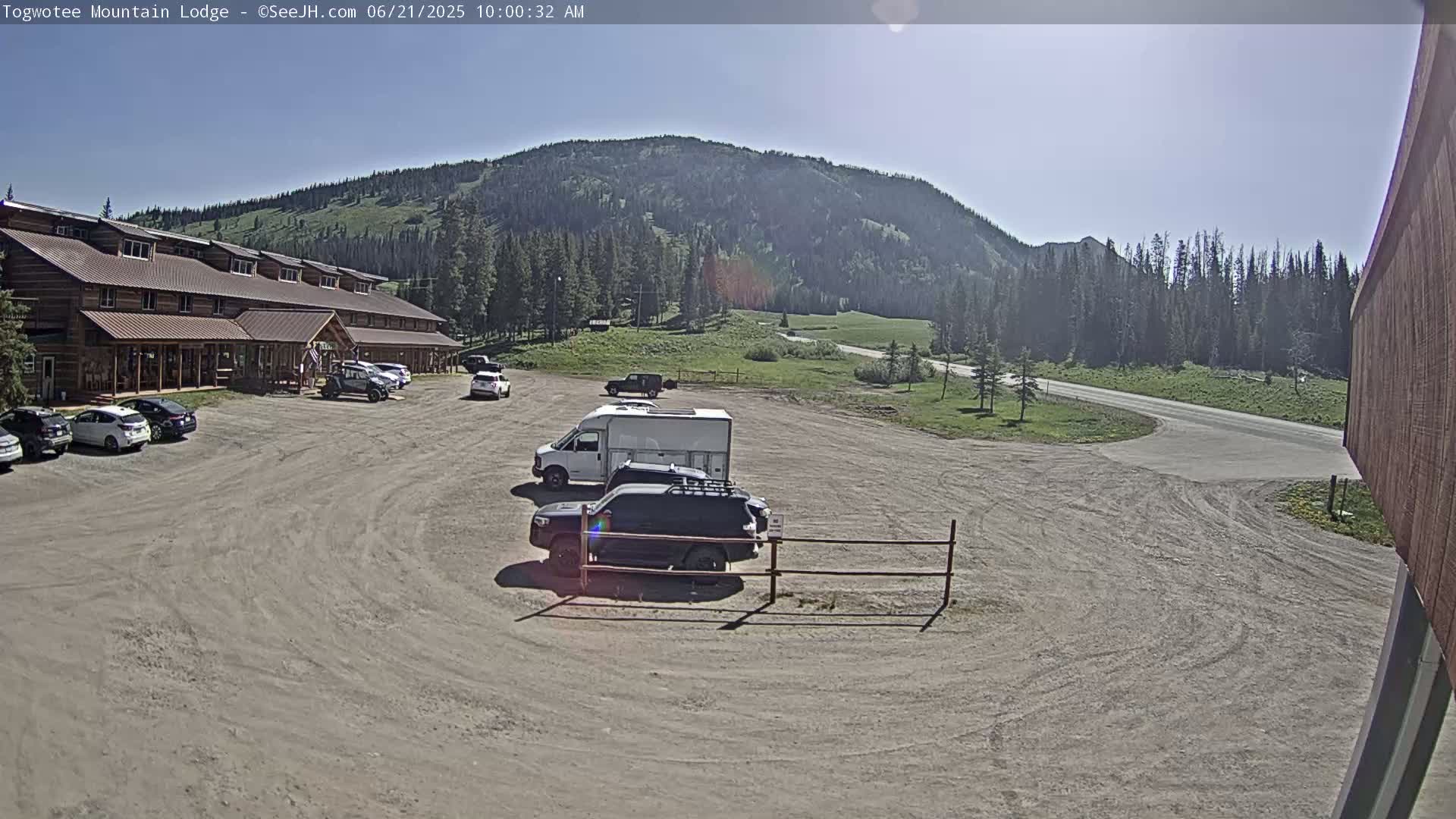 A parking lot with several cars and a van sits in front of a long, log cabin-style building, with a mountain and trees visible in the background under a clear, sunny sky.