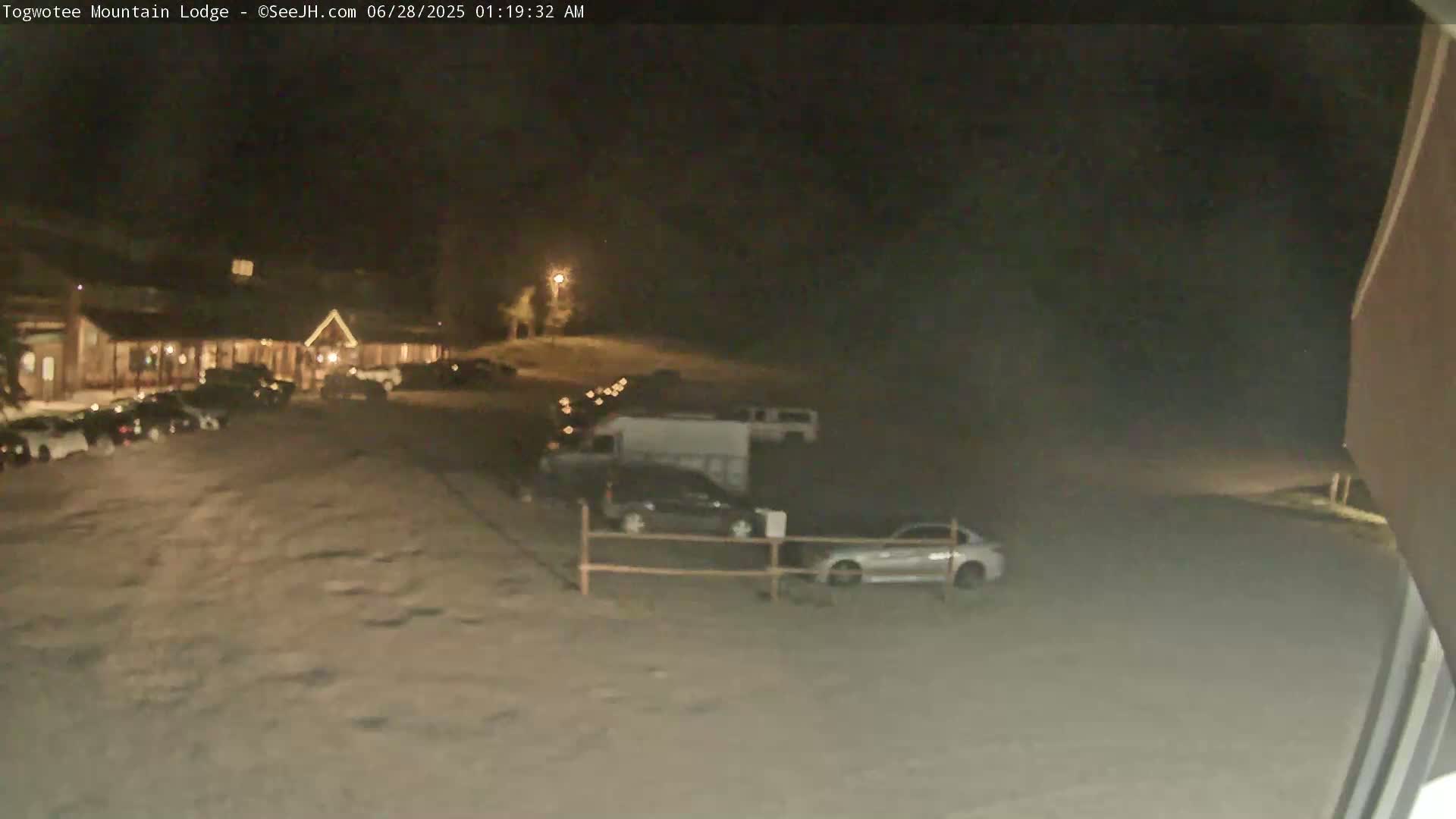 A nighttime view of a lodge parking lot with several cars and a van parked, under a dark sky.