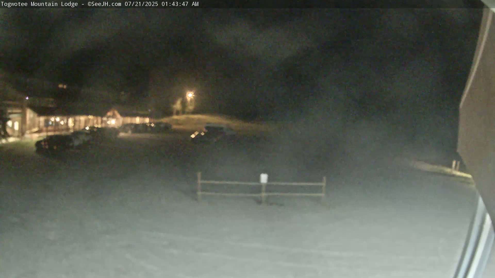 A nighttime, snowy scene shows a building with cars parked in front, a low fence, and a hazy atmosphere.
