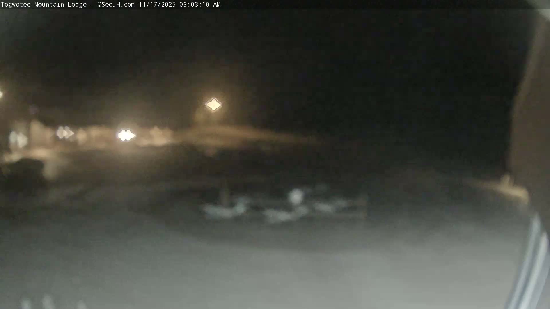 A dark and blurry outdoor night scene reveals diffuse artificial lights and indistinct shapes on the ground, indicating low visibility due to fog or heavy precipitation.