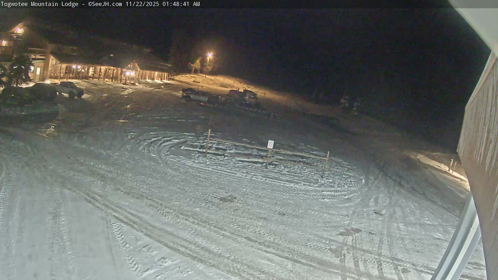 A snow-covered lodge and its surrounding parking area are illuminated at night, revealing numerous tire tracks on the ground under clear, dark conditions.
