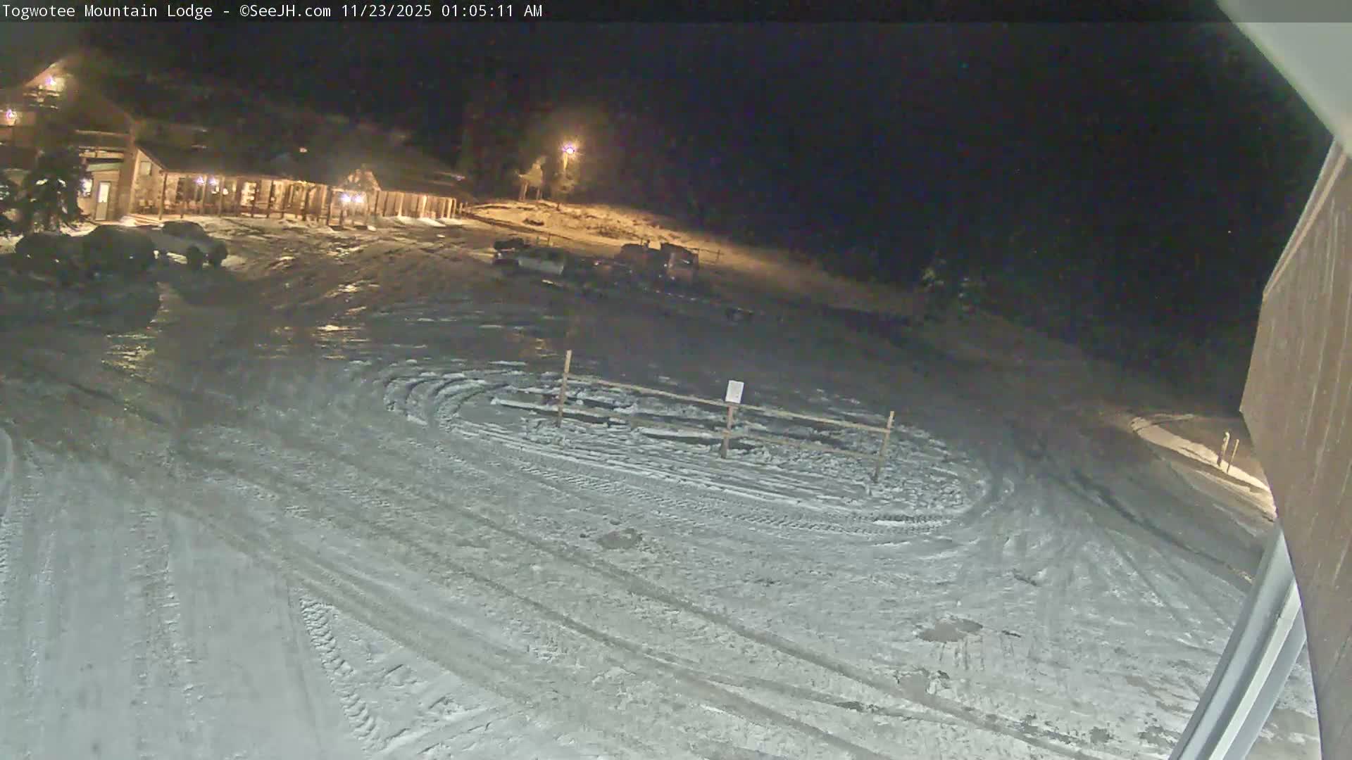 A snow-covered mountain lodge with warm lights on stands beside a parking lot marked with tire tracks, all under a clear, cold night sky.