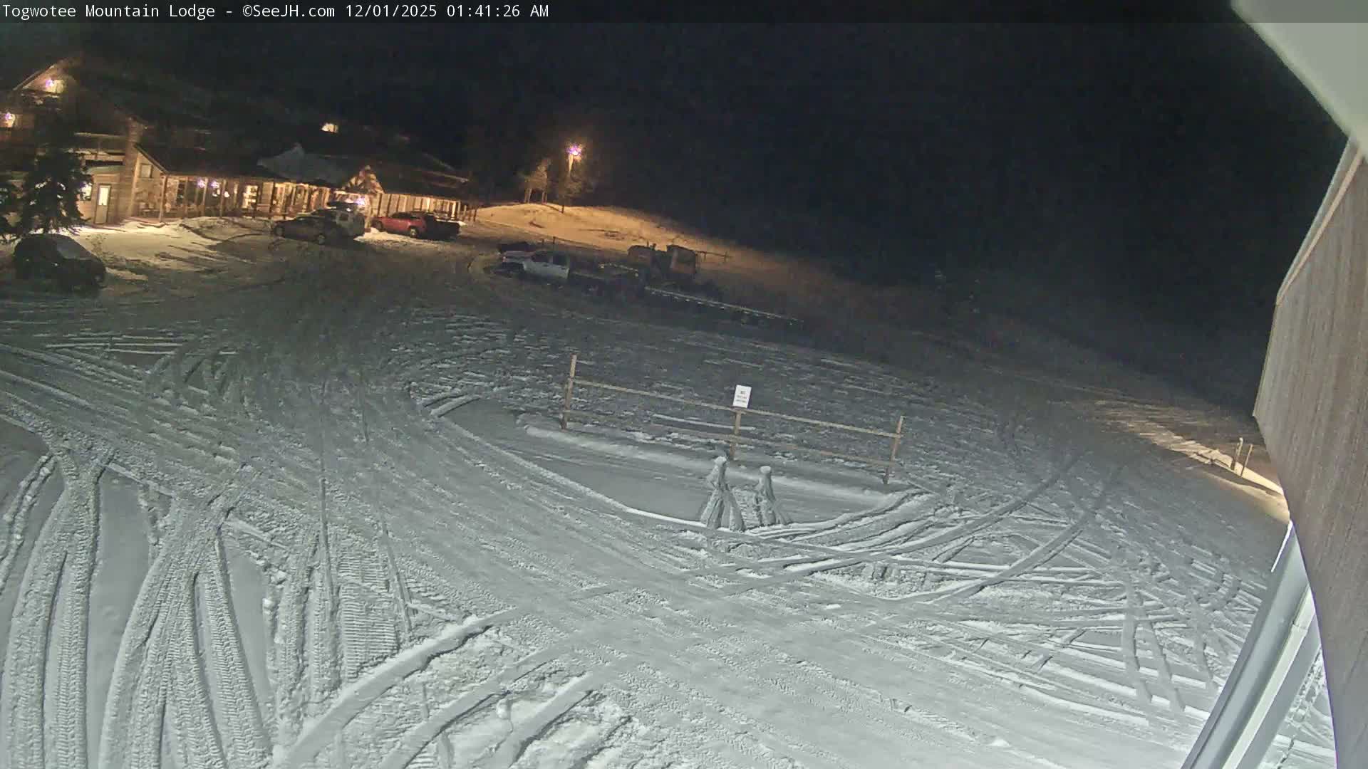 A snow-covered lodge and parking area with several vehicles and numerous tracks in the snow are illuminated at night under clear, cold conditions.