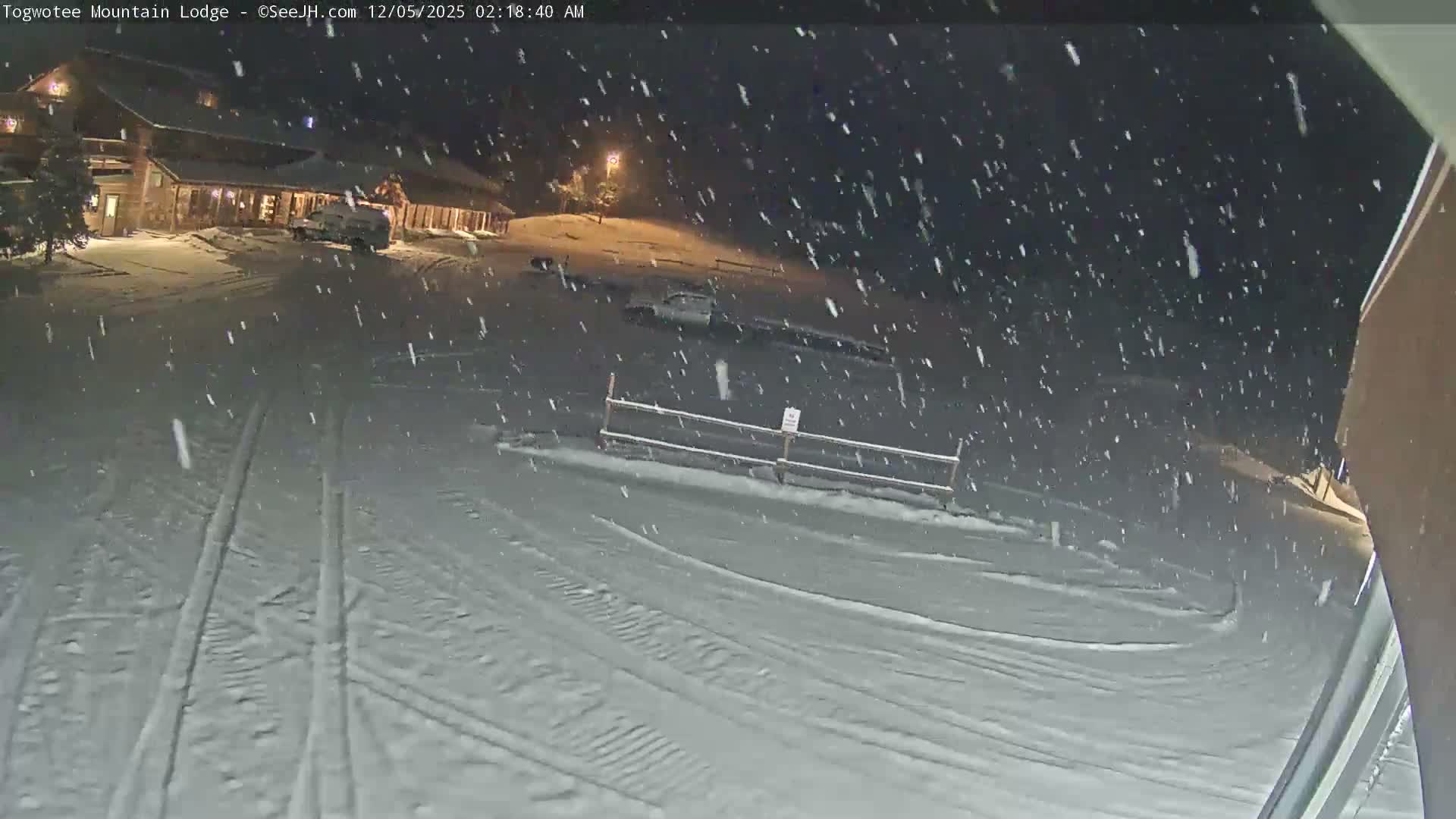 A snow-covered mountain lodge with parked vehicles and a fence is visible at night under heavy, actively falling snow.