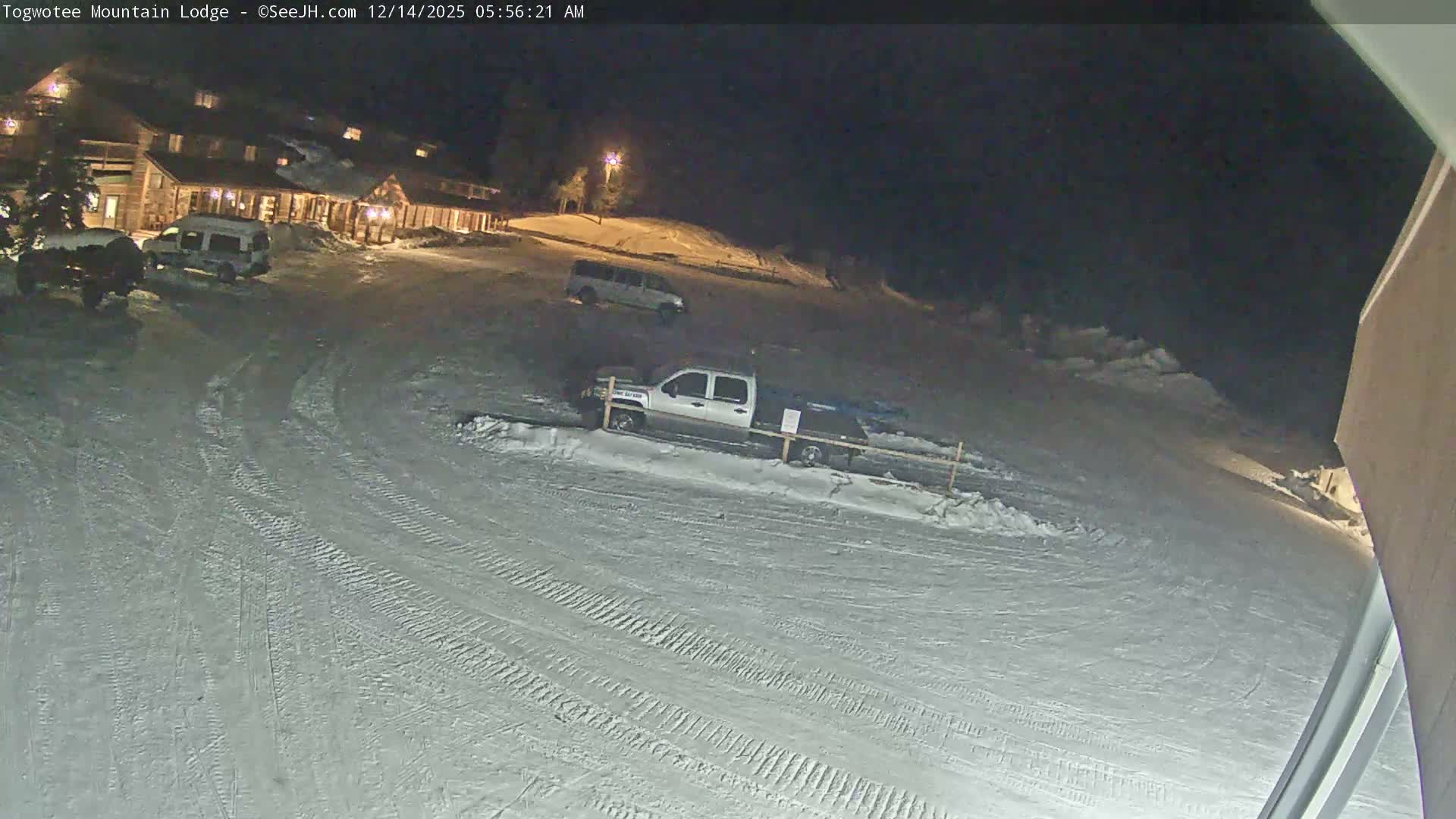 A snow-covered mountain lodge with parked vehicles and a fence is visible at night under heavy, actively falling snow.
