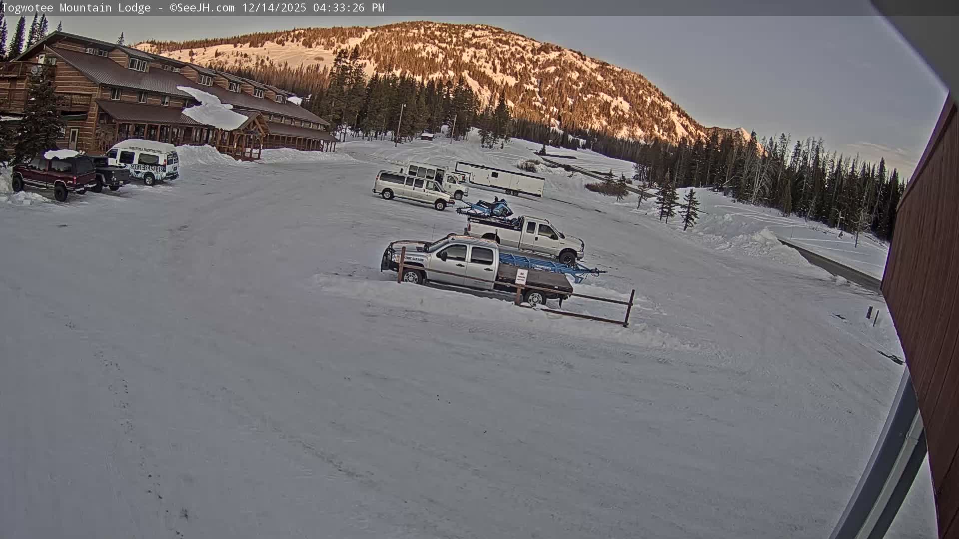 A snow-covered mountain lodge with parked vehicles and a fence is visible at night under heavy, actively falling snow.