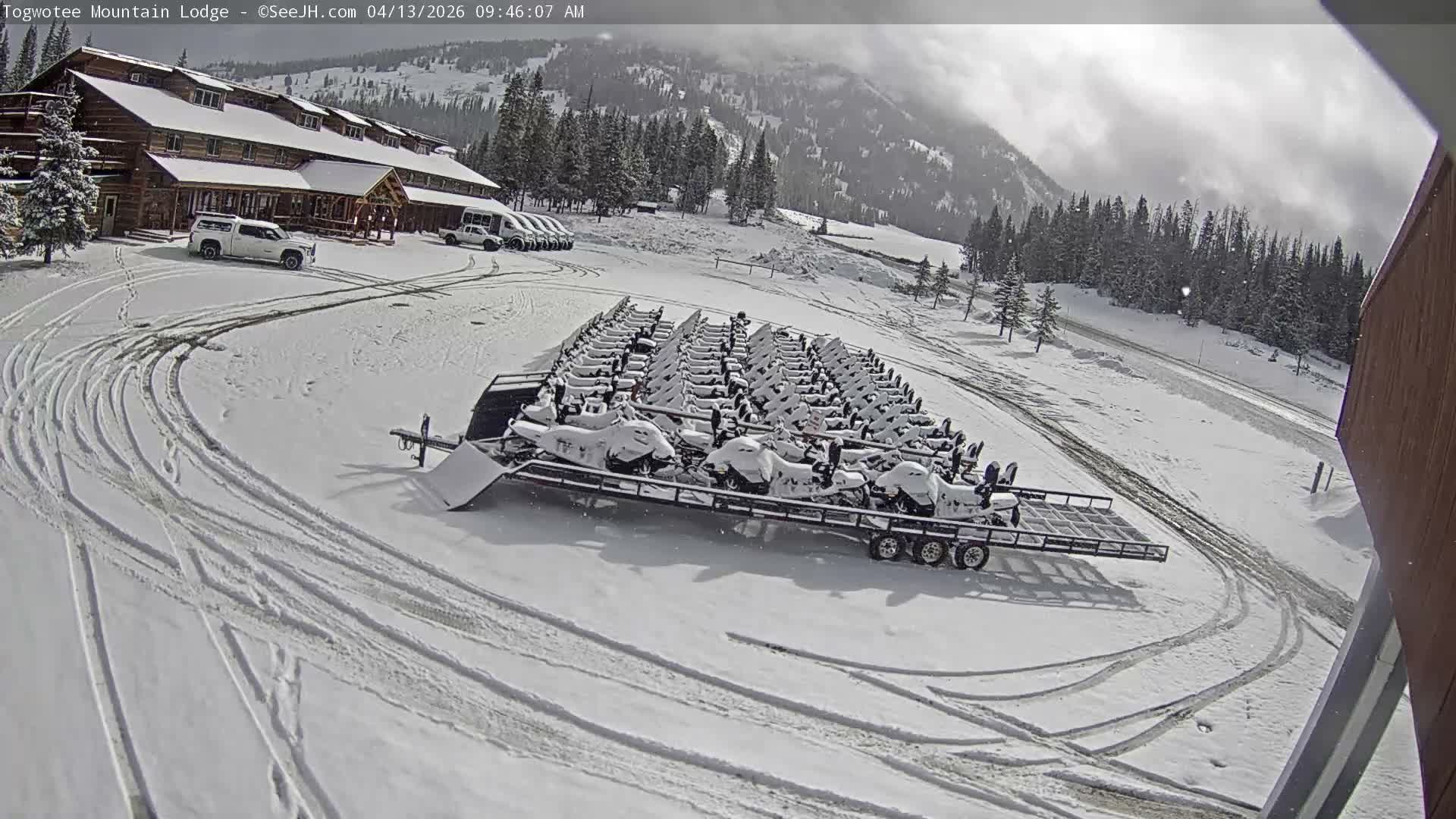 A snow-covered mountain lodge with parked vehicles and a fence is visible at night under heavy, actively falling snow.