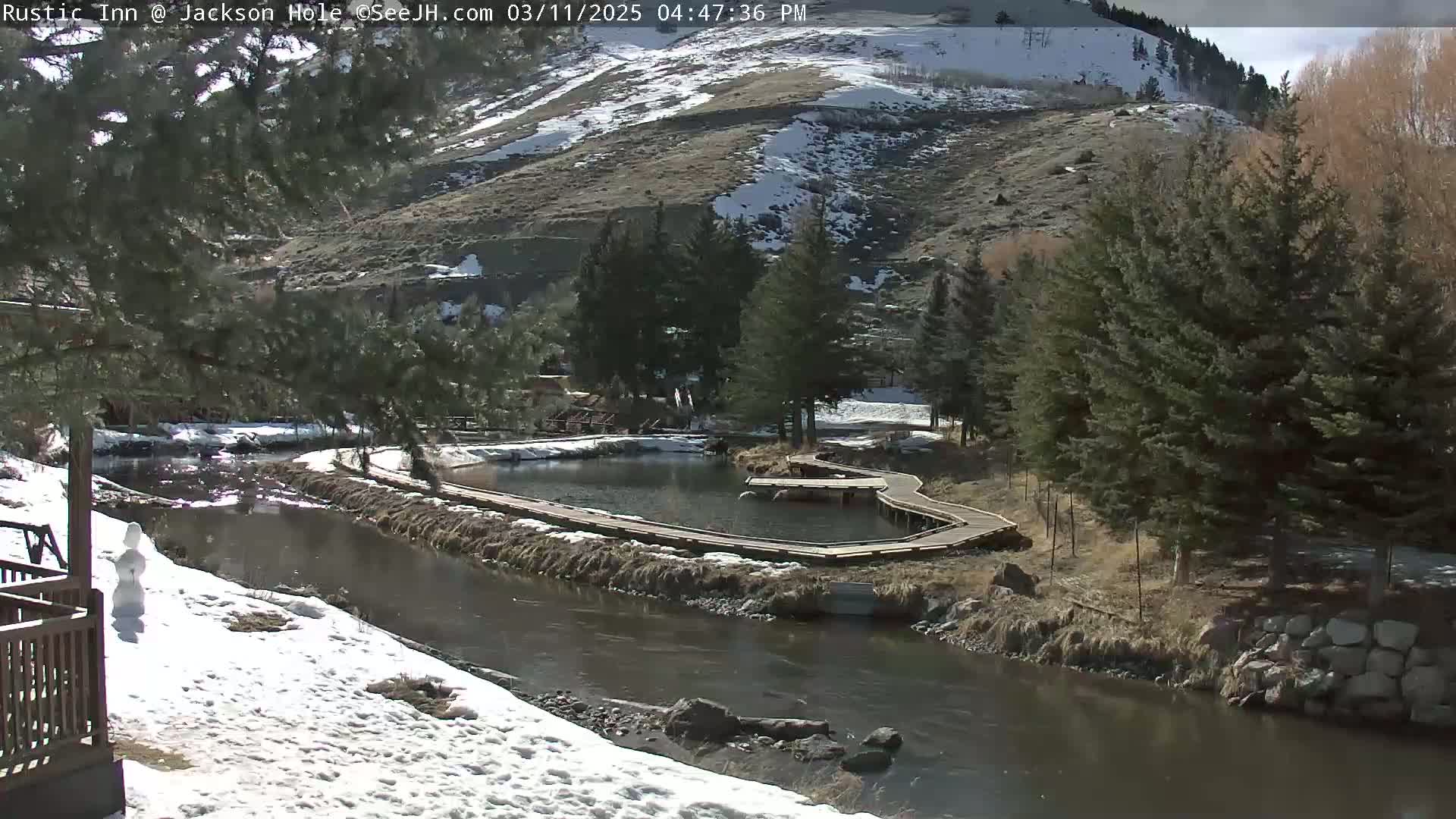 A sunny day shows a river flowing past a snow-dusted landscape with wooden walkways around a small pond next to evergreen trees and a snow-capped hill in the background.