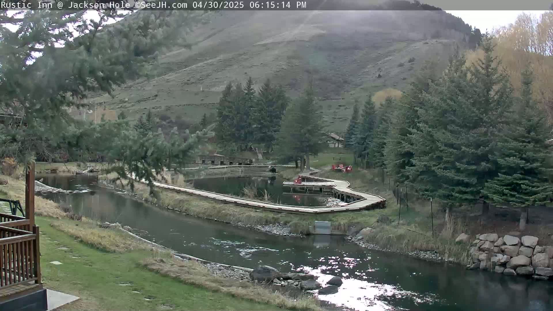 A calm river flows through a green valley, alongside wooden walkways and evergreen trees under a partly cloudy sky.