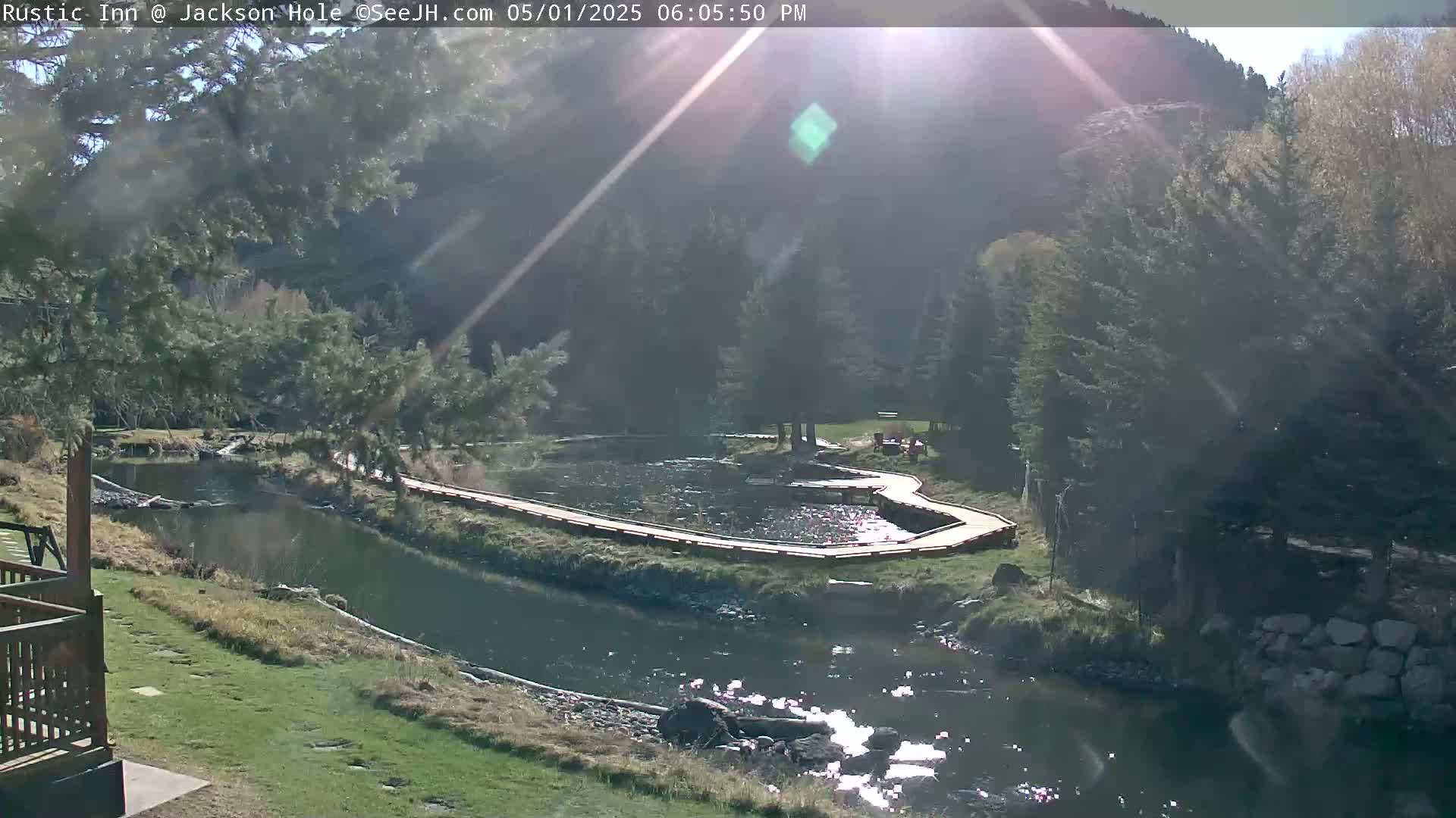 A sunny day reveals a river flowing through a green valley, with a winding wooden walkway alongside a calm section of the water, surrounded by evergreens and hills.
