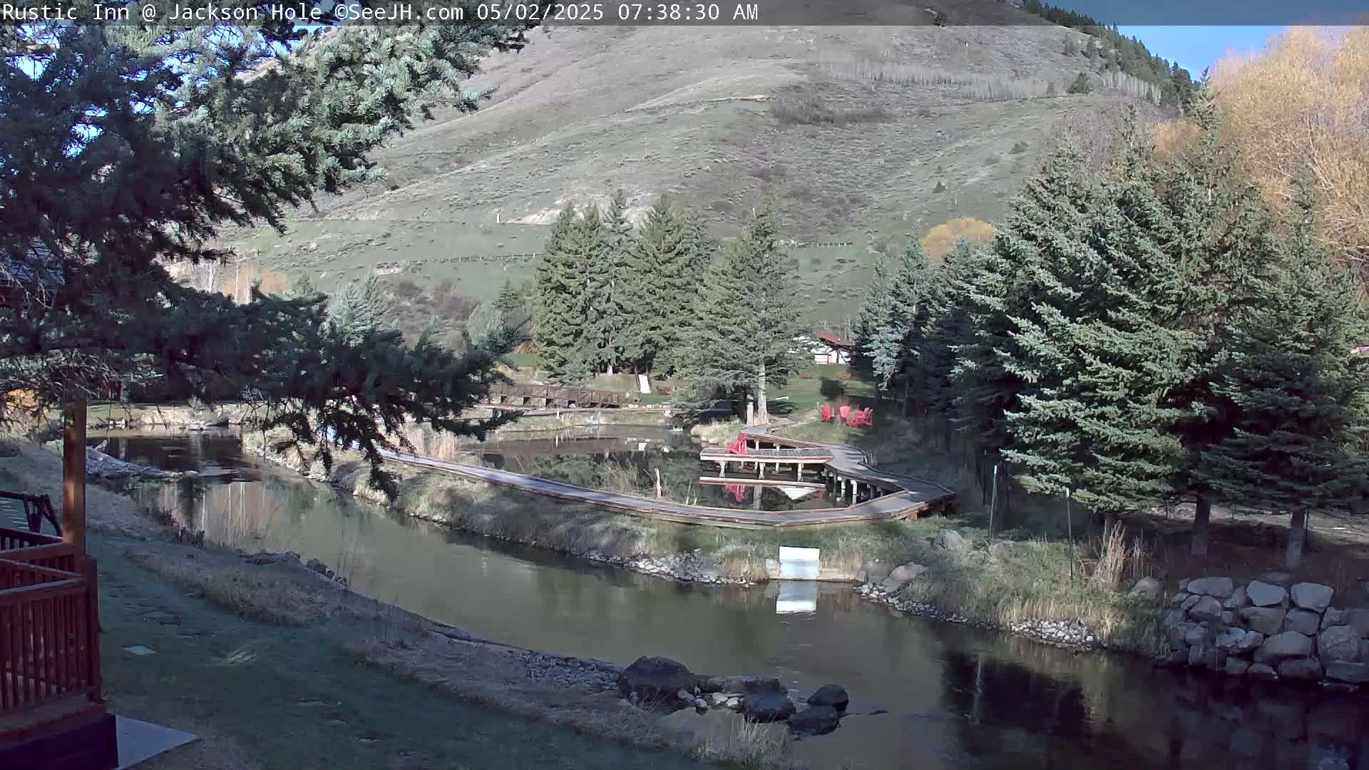 A sunny view of a calm river winding through a wooded area with boardwalks and small ponds, nestled against a hillside.