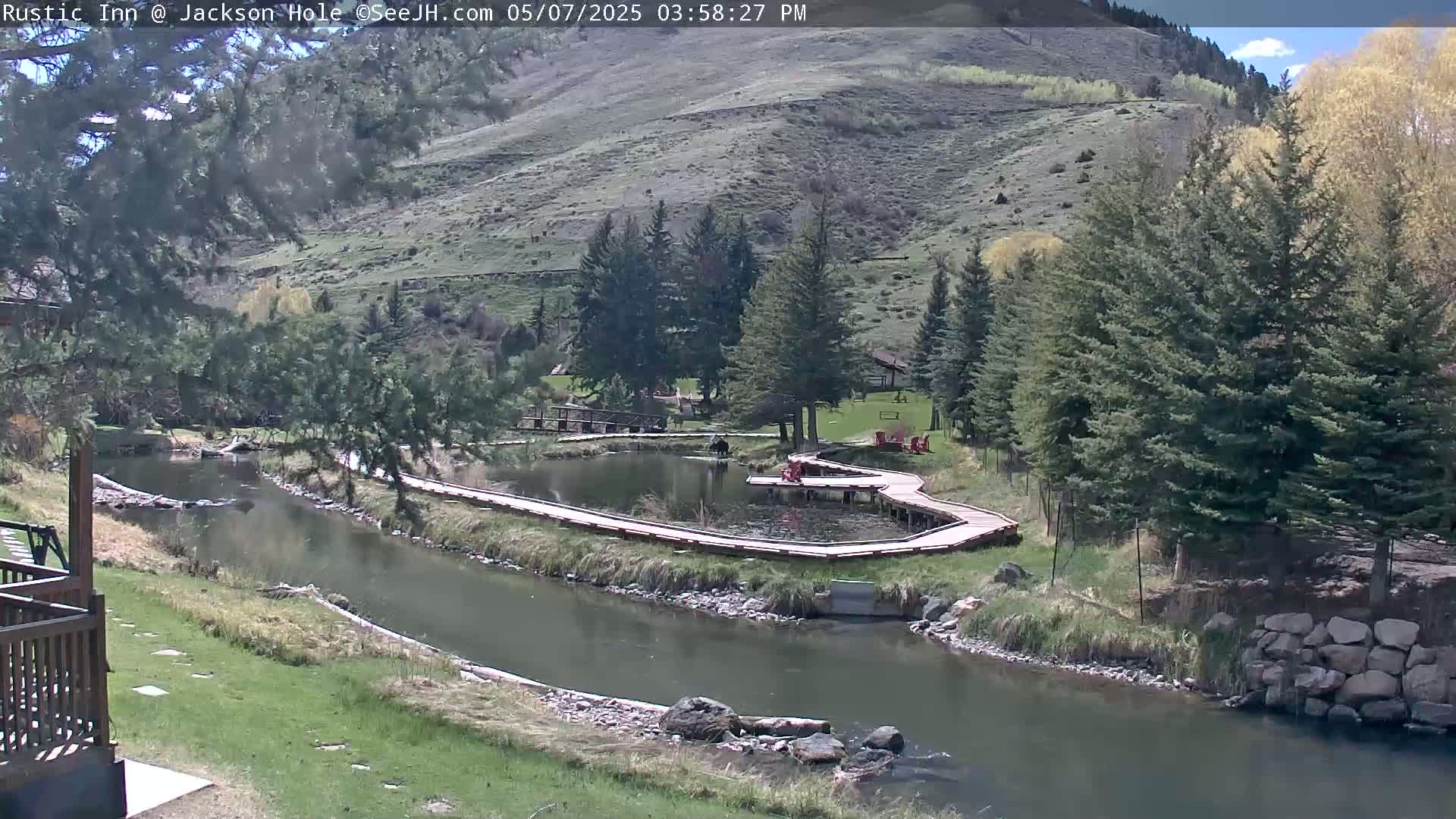 A river flows through a green valley alongside a wooden boardwalk that curves around a pond, all under a partly sunny sky.