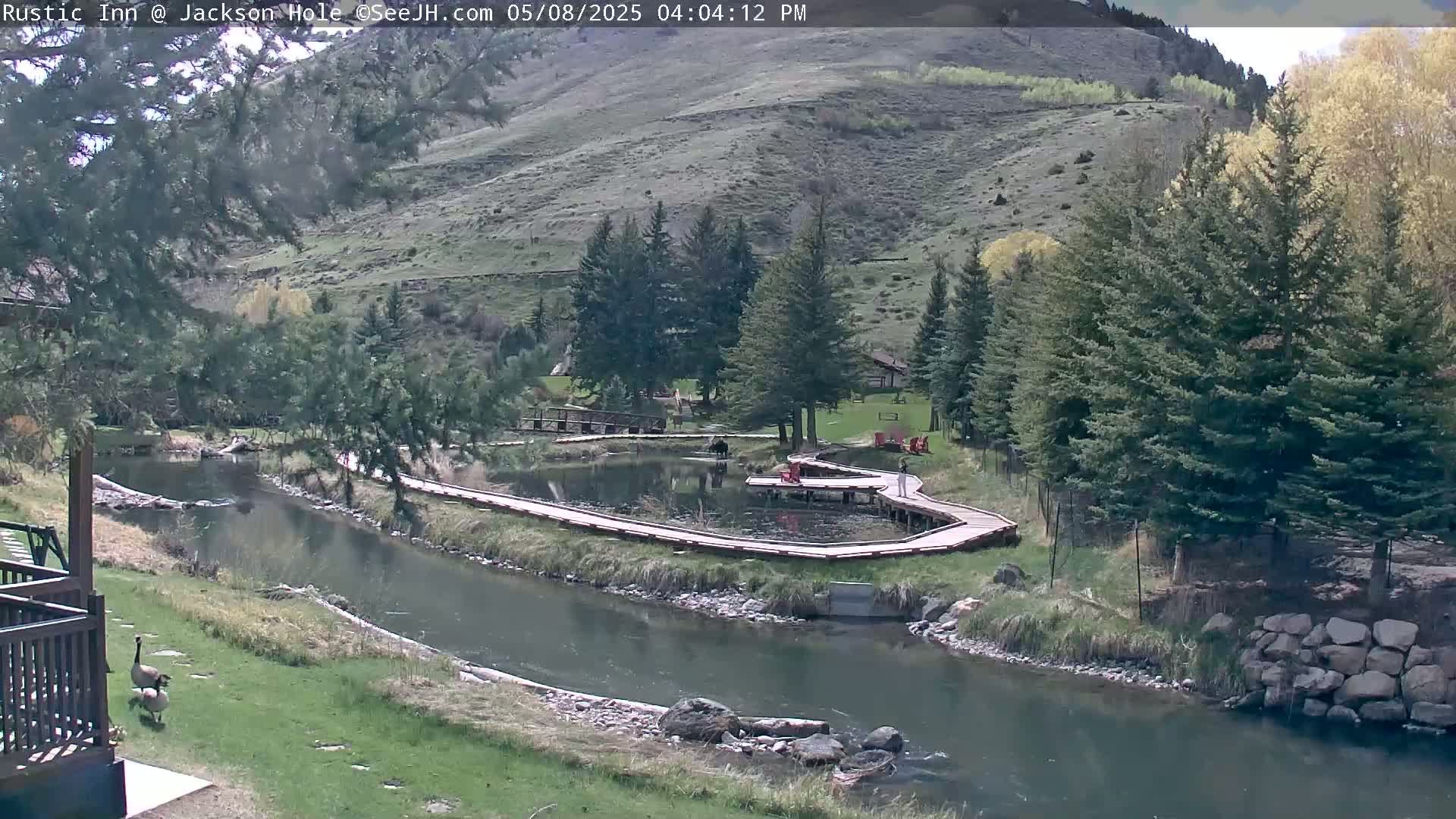 A tranquil river flows through a green valley, alongside wooden walkways and a small pond, under a partly sunny sky.
