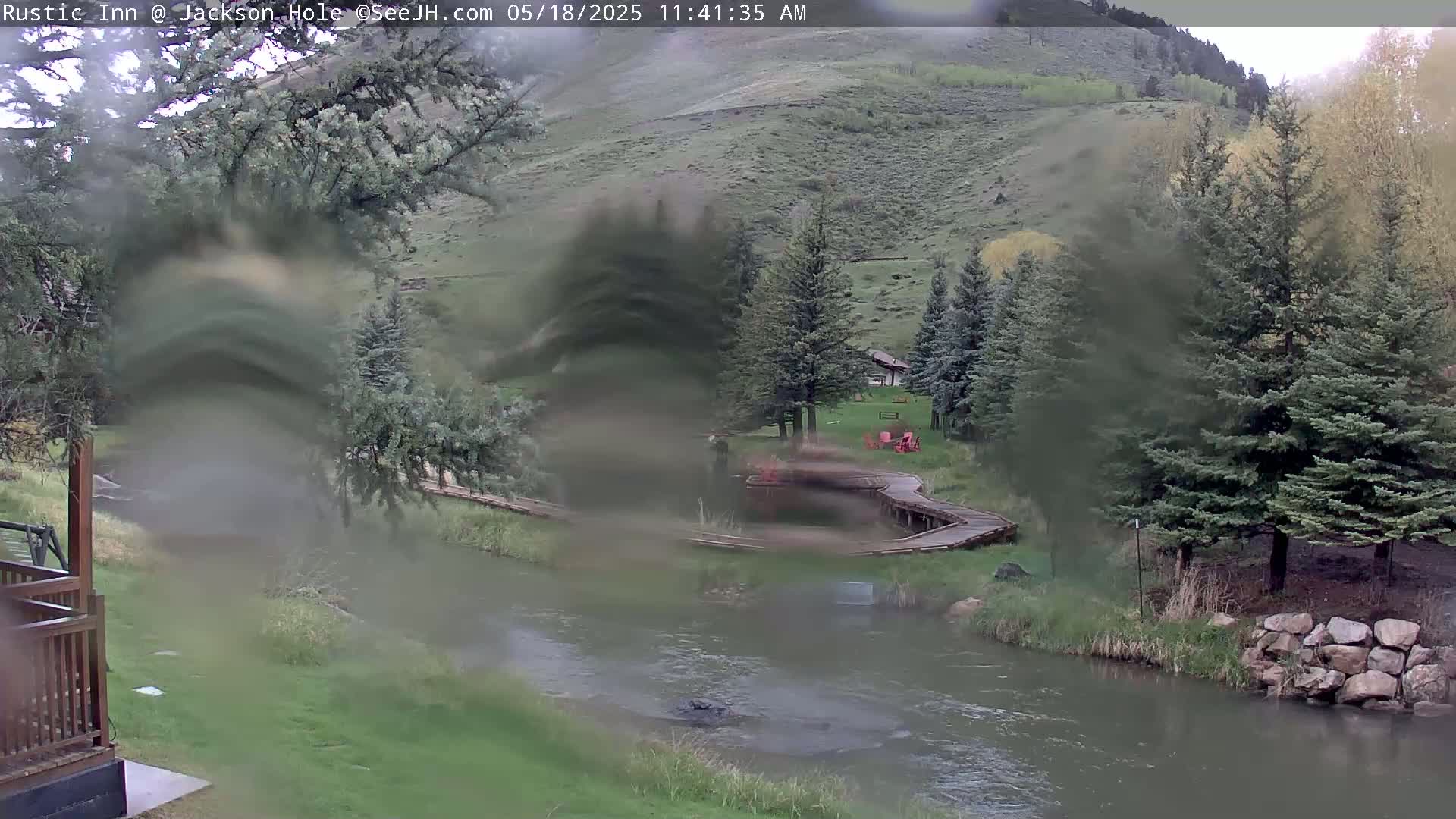 A rain-blurred view of a river flowing past a grassy area with wooden walkways and evergreen trees, set against a hillside.