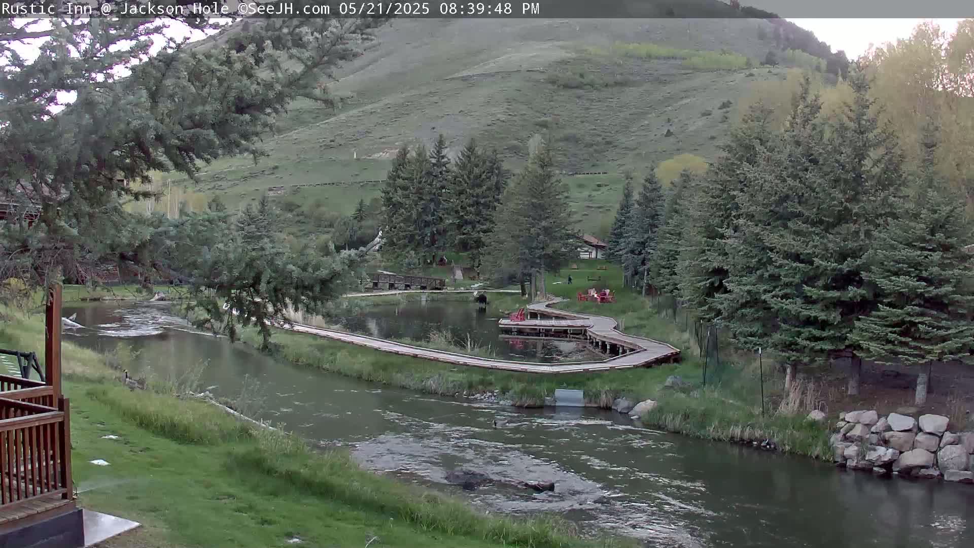 A calm river flows past a wooden boardwalk surrounding a pond, nestled in a valley with lush green hills and evergreen trees under a partly cloudy sky.