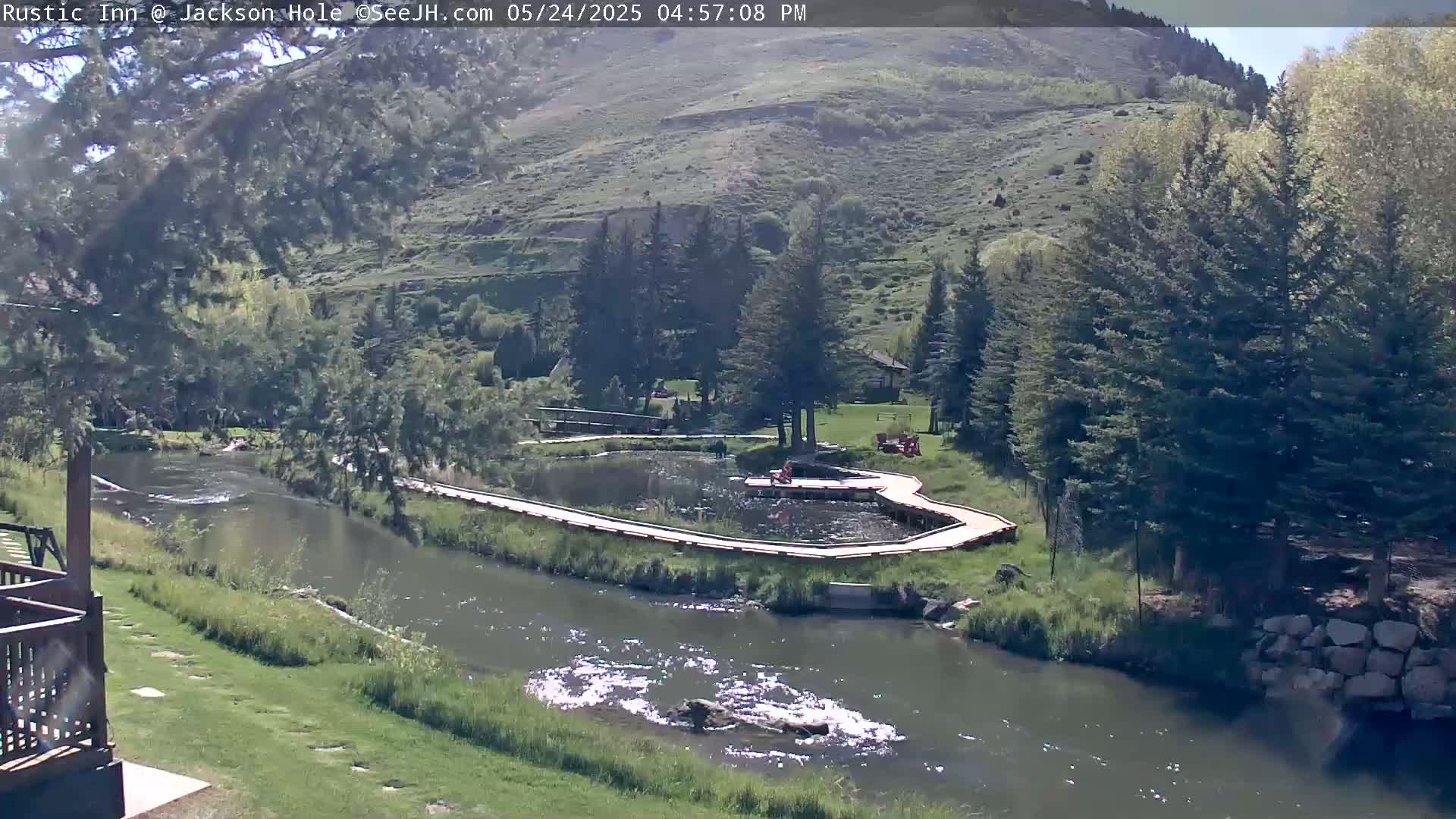A river flows past a wooden walkway surrounding a small pond, nestled in a valley with green hills and trees under a sunny sky.
