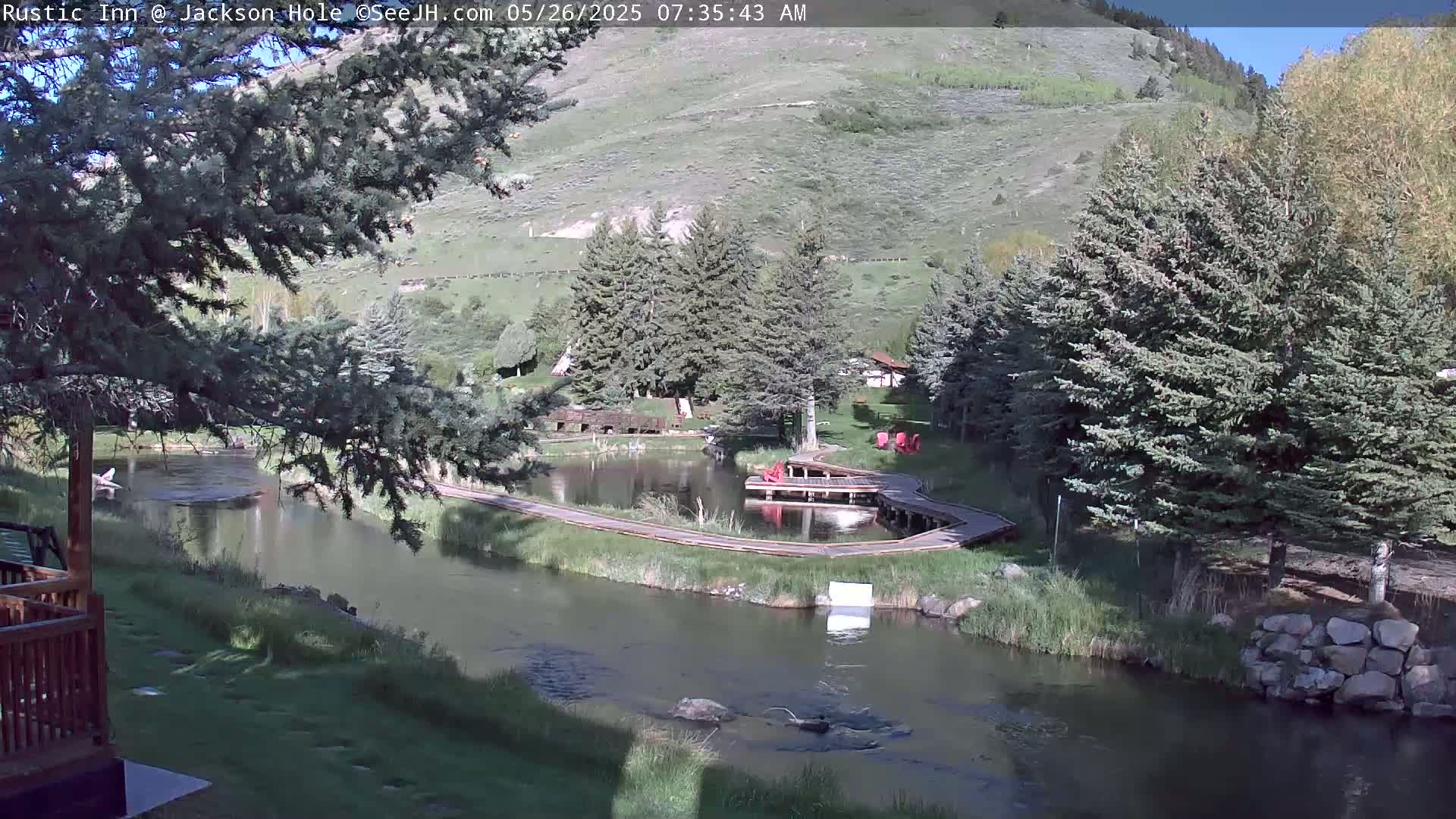 A calm river flows through a green valley under a sunny sky, past a pond with wooden walkways and several evergreen trees.