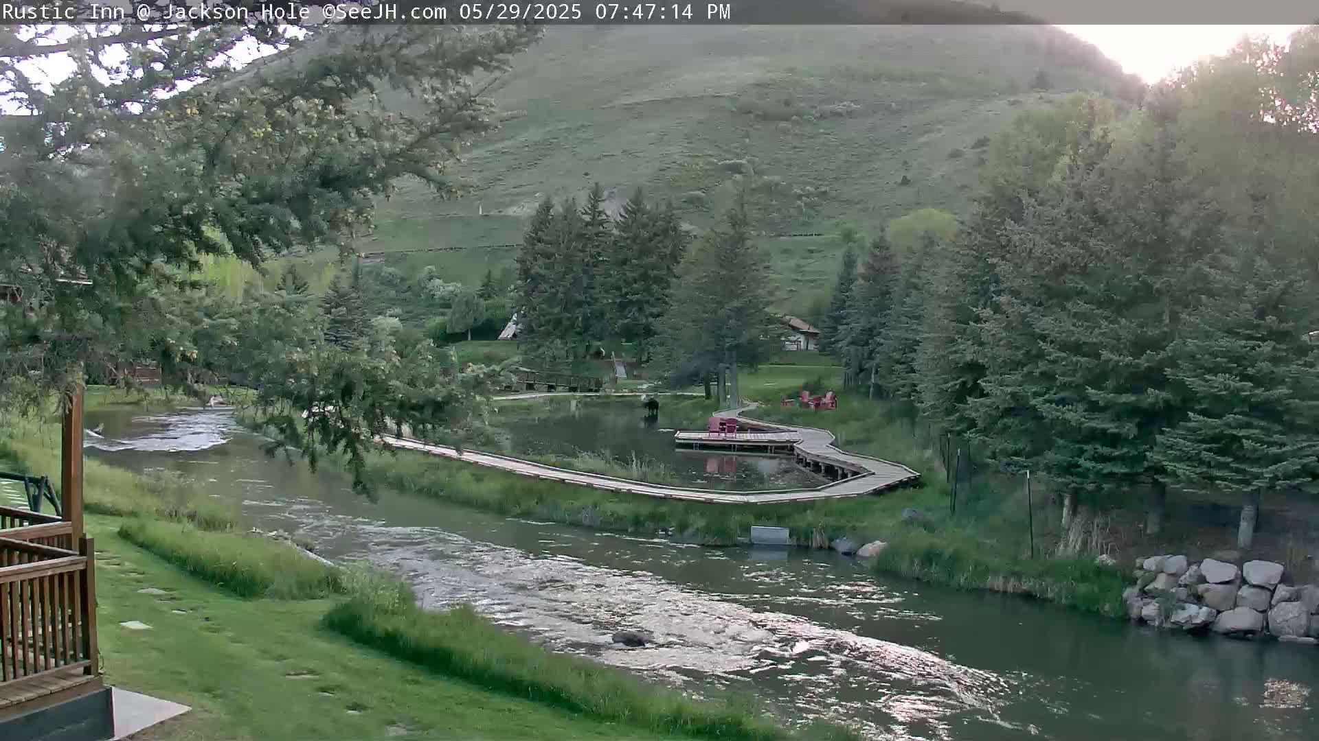 A river flows through a lush green valley under a partly sunny sky, alongside a wooden boardwalk winding near a small pond and some trees.