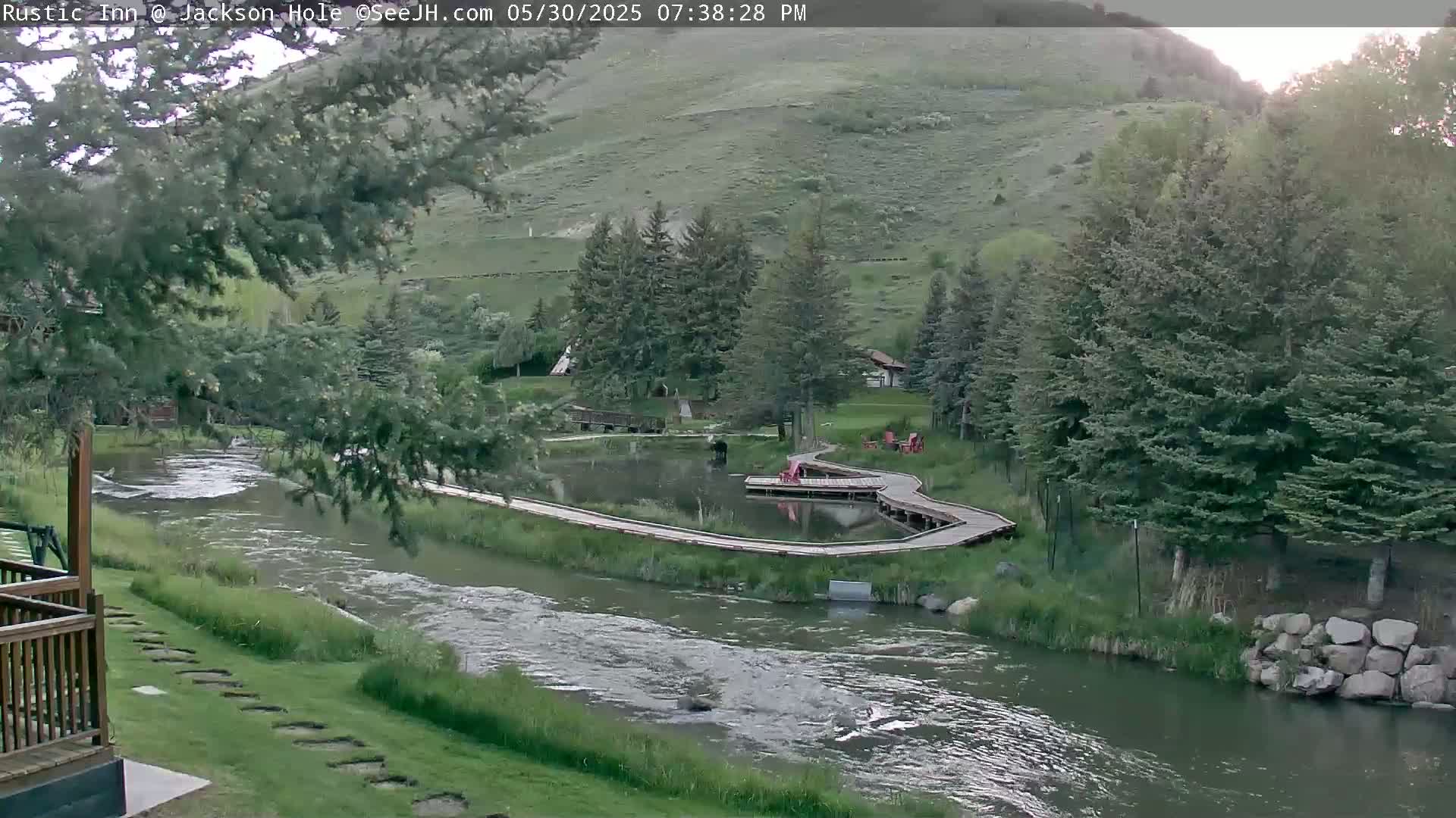 A river flows past a grassy area with a wooden boardwalk surrounding a pond, nestled among evergreen trees on a hillside under a partly cloudy sky.