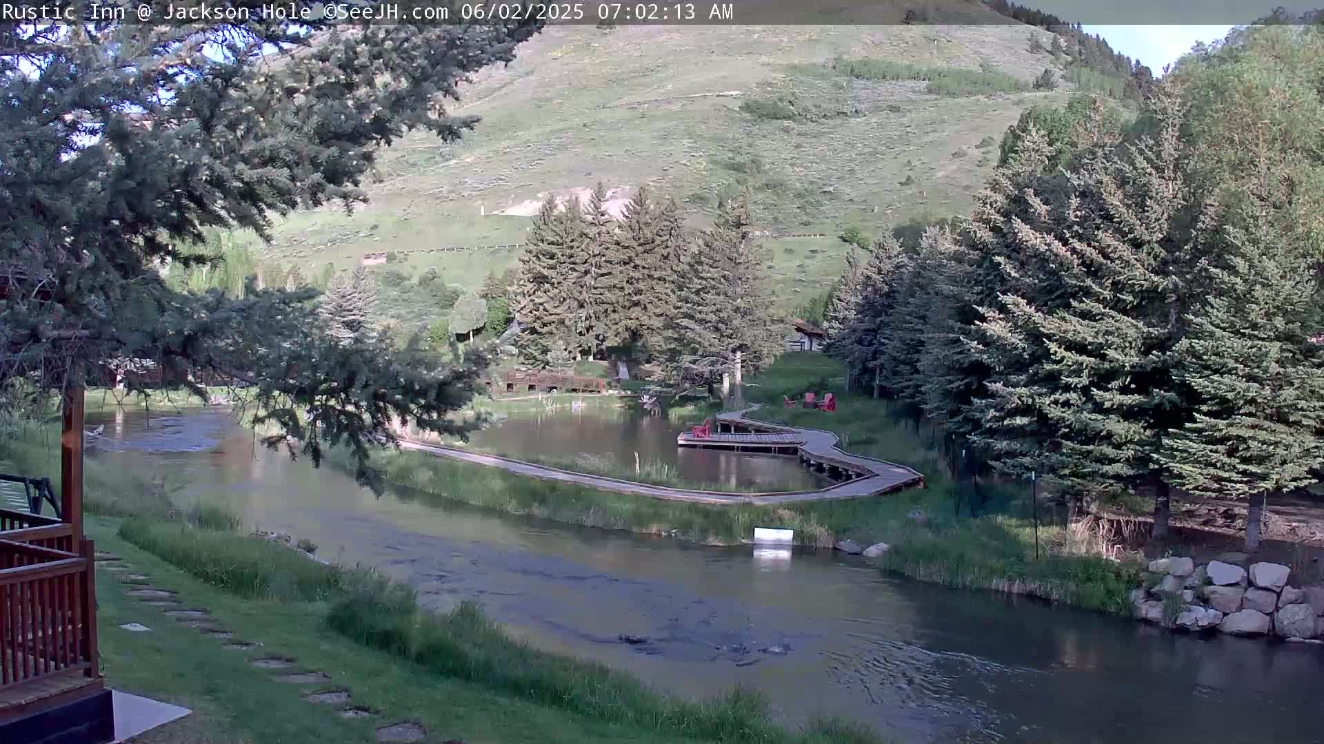 A tranquil river flows past a grassy area with a small pond and wooden walkways, all nestled amidst lush green trees and hills under a partly sunny sky.