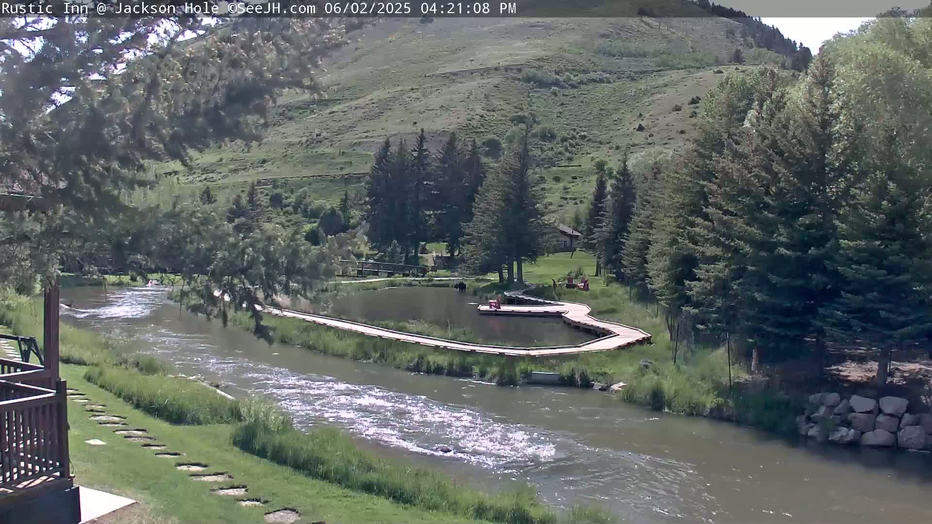 A flowing river is seen from a high vantage point on a sunny day, with a wooden walkway curving alongside a small pond and lush green hills in the background.