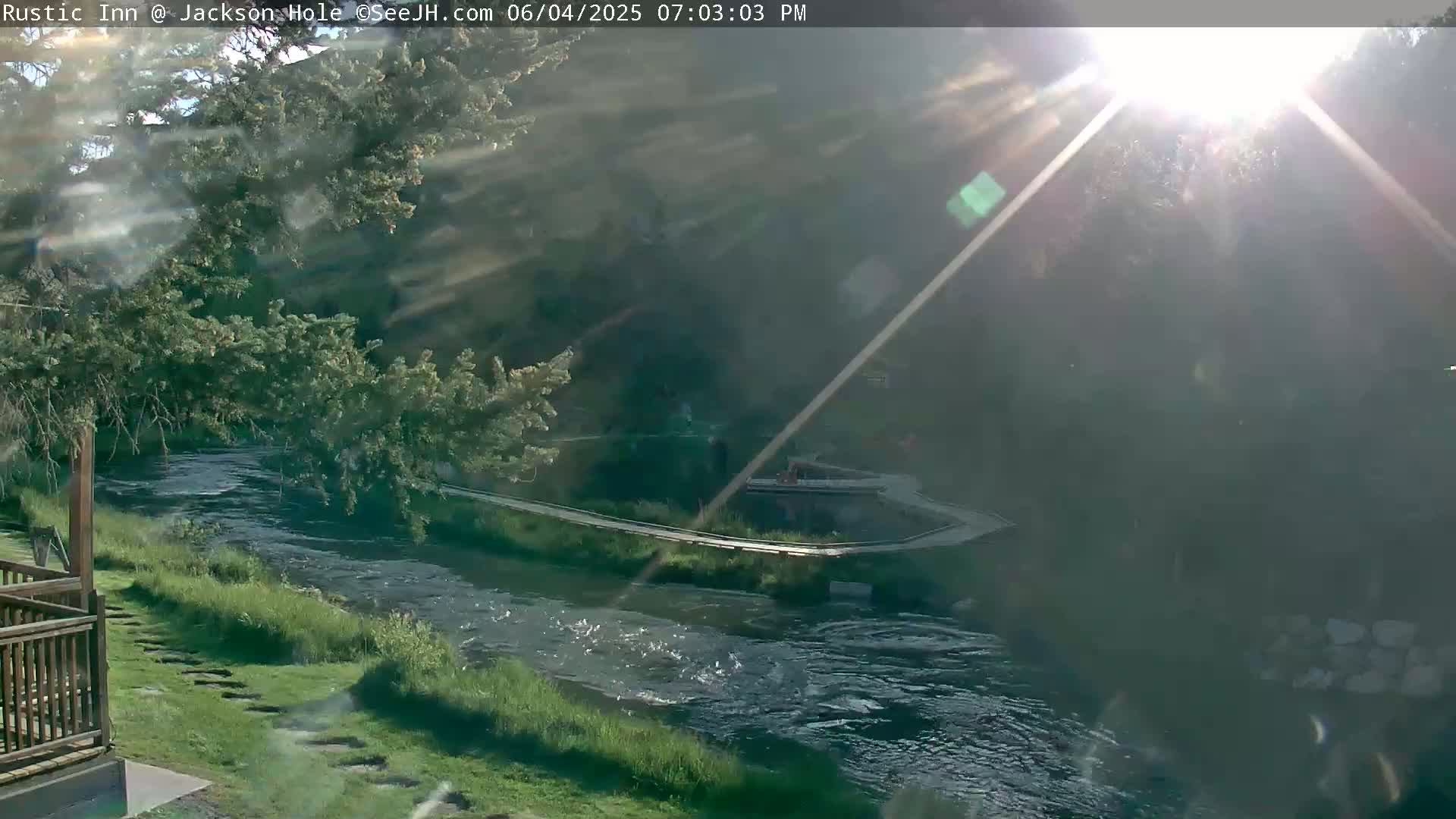 A sunny day reveals a flowing river with a wooden walkway and surrounding green vegetation.