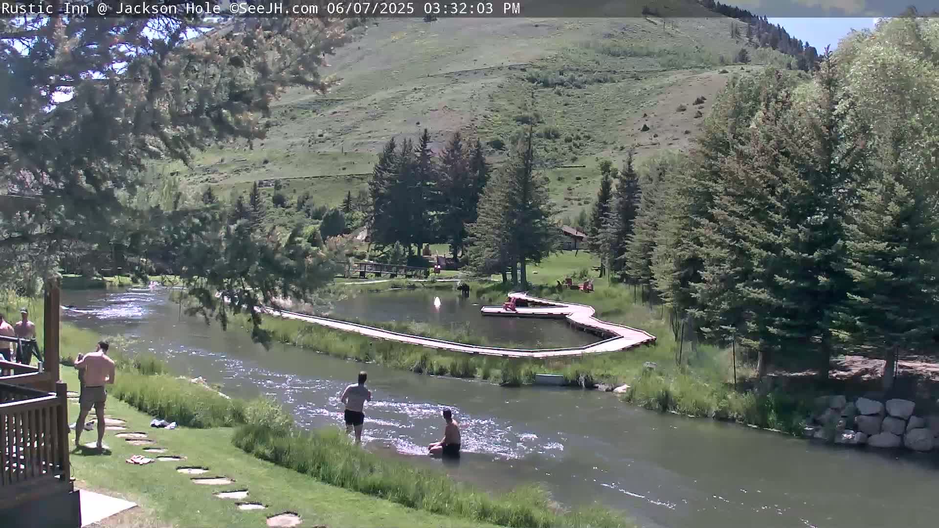A sunny day shows several people swimming and relaxing near a river that winds through a green, tree-lined valley.