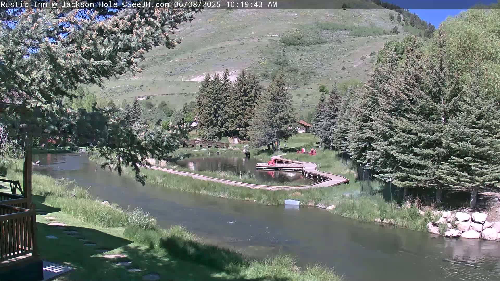 A calm river flows through a green valley under a sunny sky, past a wooden boardwalk leading to a small pond and some buildings nestled among evergreen trees on a hillside.