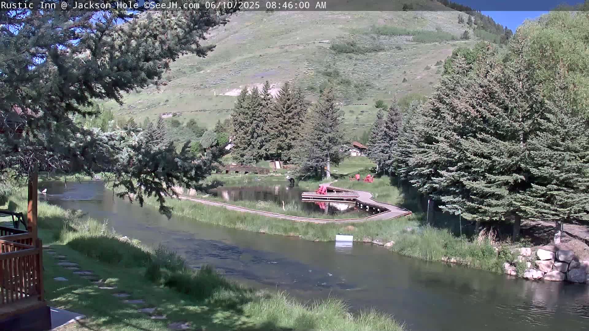 A sunny day reveals a tranquil river flowing past a wooden boardwalk and a small pond, nestled within a lush green valley surrounded by evergreen trees and hills.