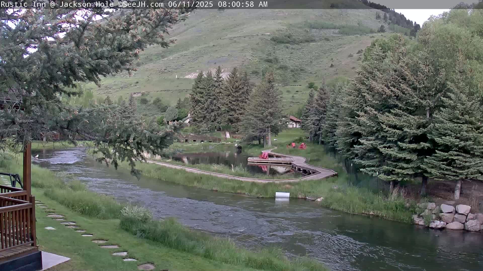A river flows past a wooden boardwalk and small pond, nestled among evergreens on a hillside under a partly cloudy sky.