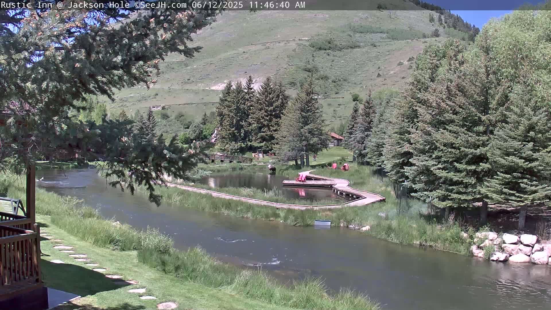A tranquil river flows past a grassy area with wooden walkways and a small pond, all nestled amongst lush green trees and hills under a sunny sky.