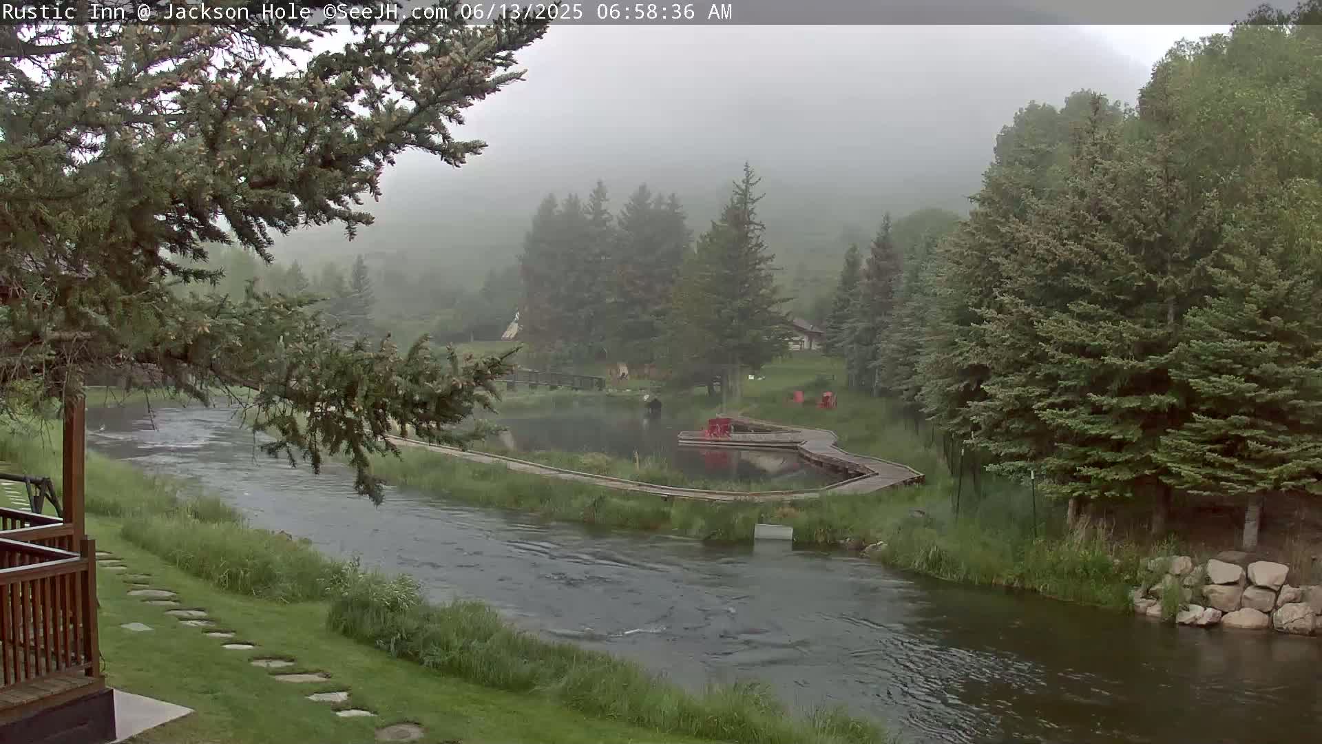 A foggy morning scene shows a river flowing past a wooden walkway and small pond, surrounded by evergreen trees and grassy areas.