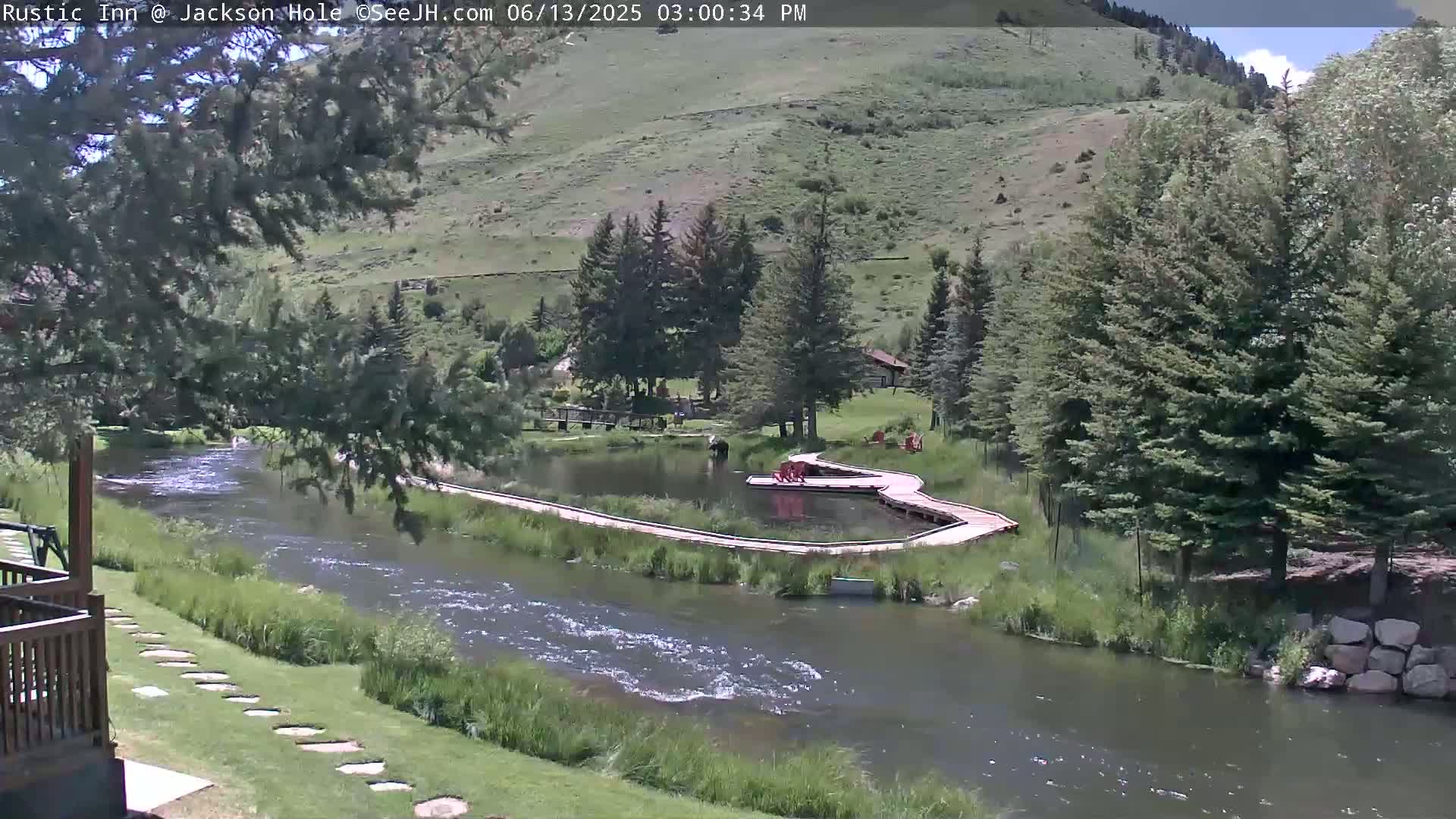 A river flows past a wooden boardwalk and small pond, surrounded by lush green vegetation and a hill in the background under a partly sunny sky.