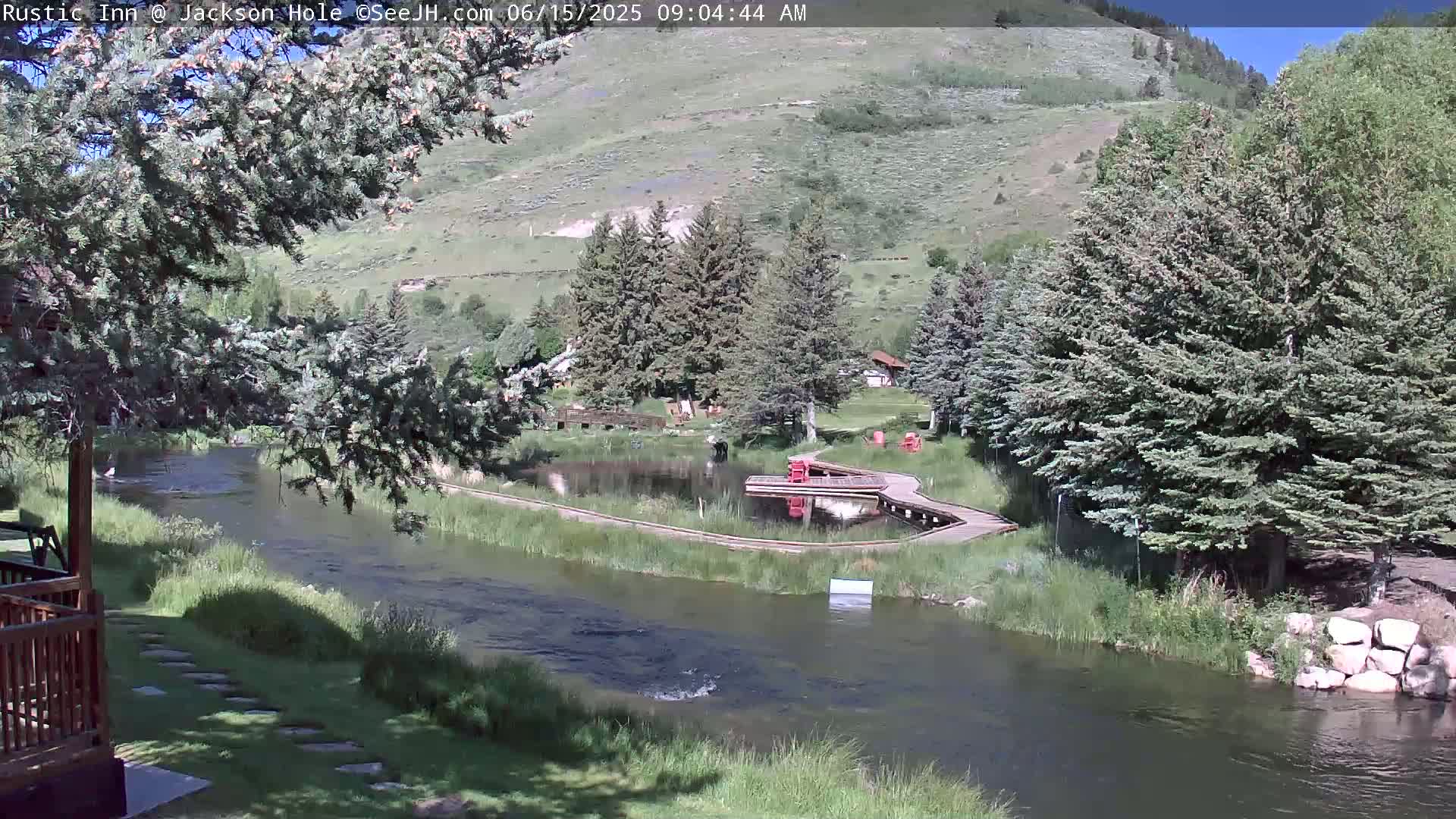 A river flows past a small pond with a wooden walkway and red chairs, surrounded by green trees and hills under a sunny sky.