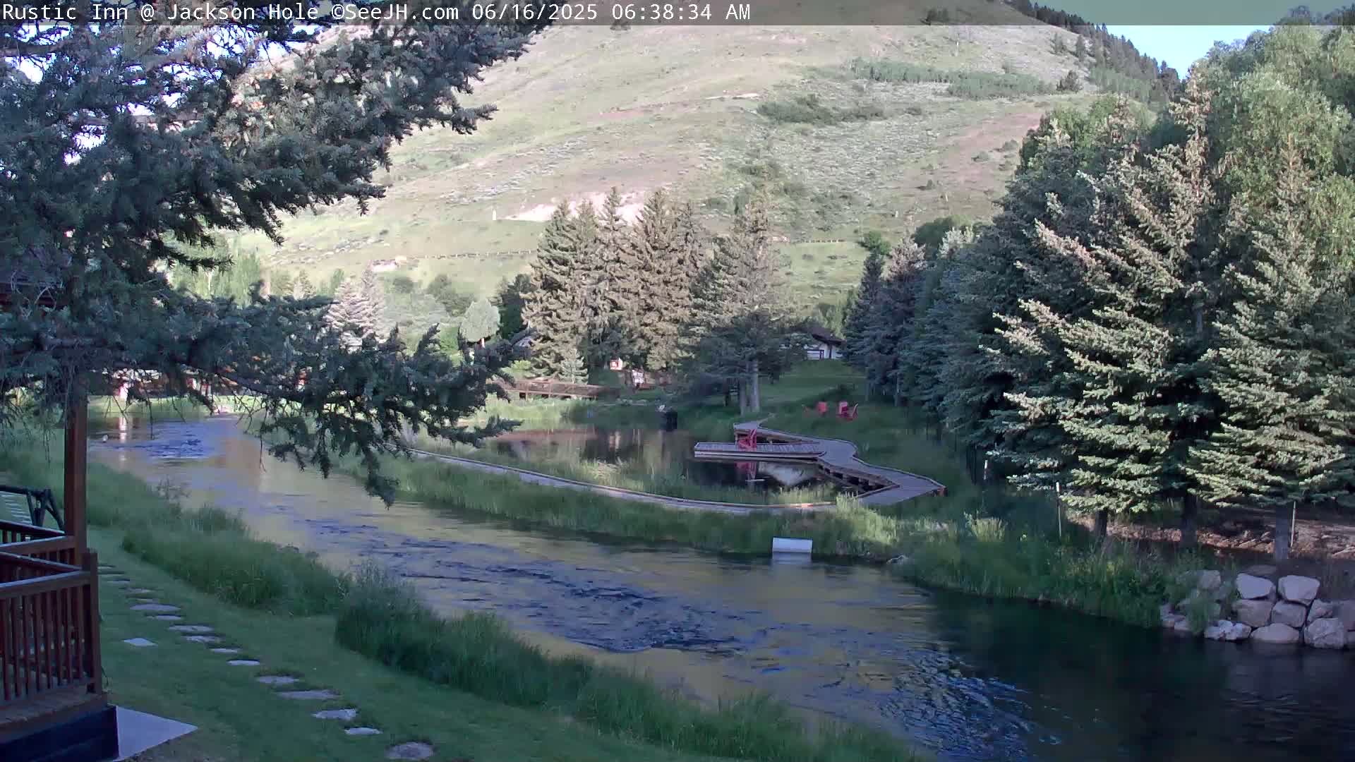 A calm river flows through a green valley, bordered by evergreen trees and wooden walkways under a clear sky.