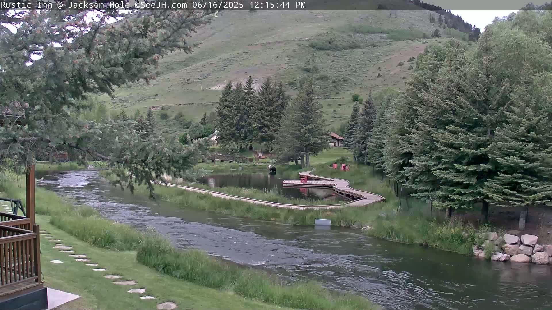 A river flows past a grassy area with a pond and wooden walkways, all nestled against a hillside covered in green trees under a partly cloudy sky.
