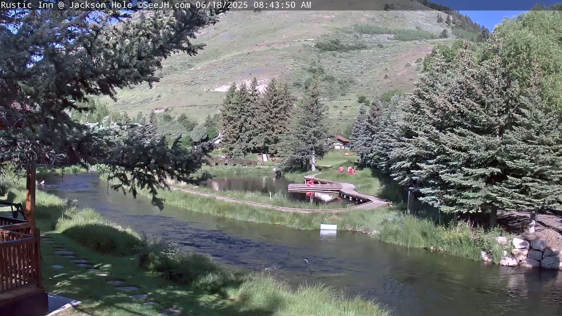 A river flows through a sunny green valley with trees, a small pond, and a wooden walkway leading to a dock.