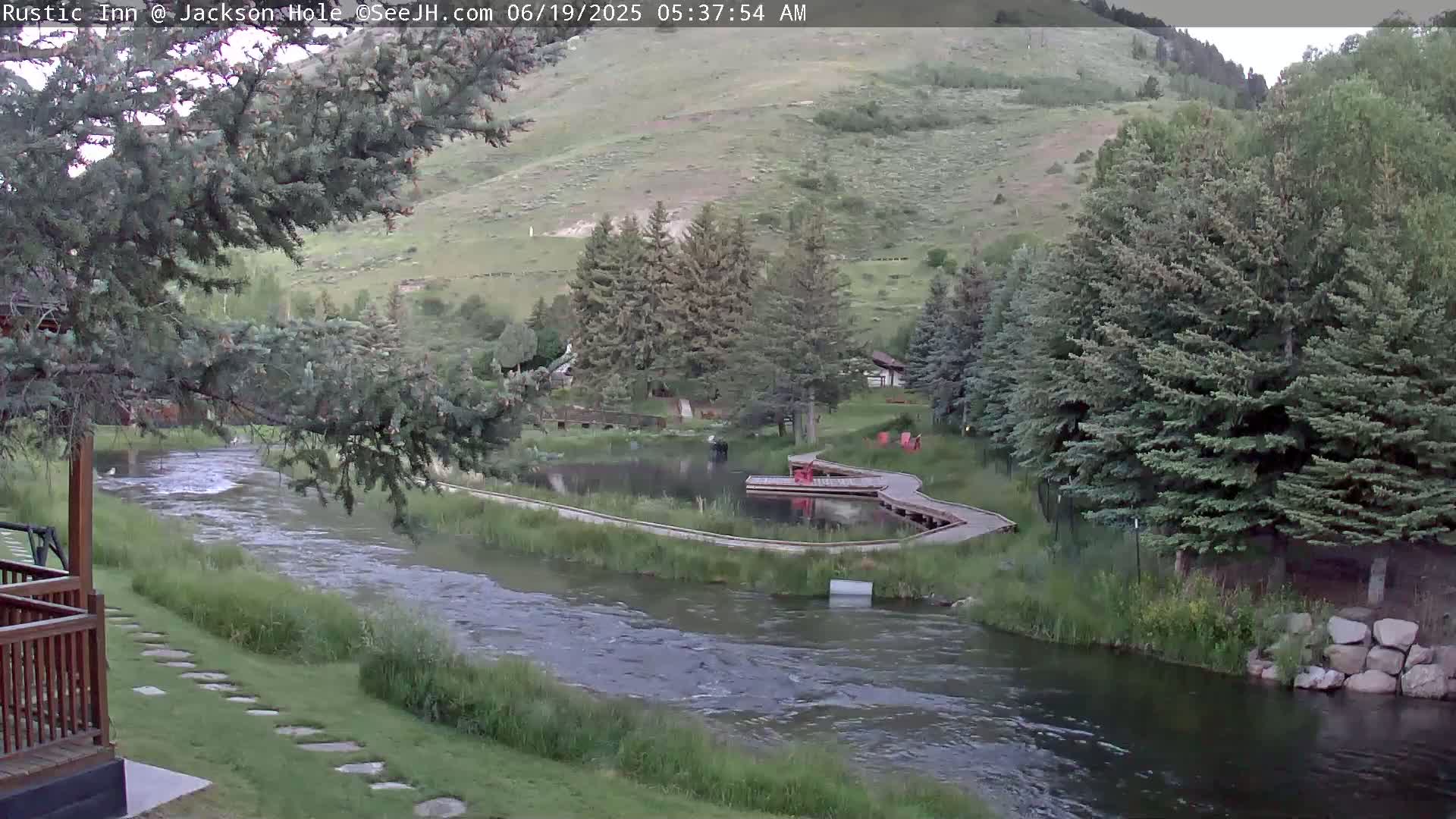 A flowing river curves through a green landscape dotted with evergreen trees and a small pond, under a clear sky.