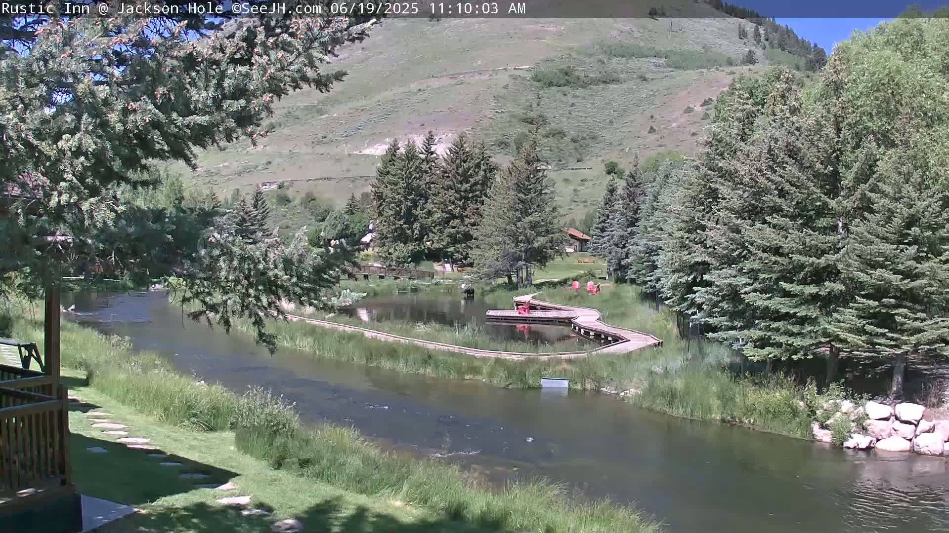 A calm river flows through a green valley under a sunny sky, alongside a wooden boardwalk that encircles a small pond.