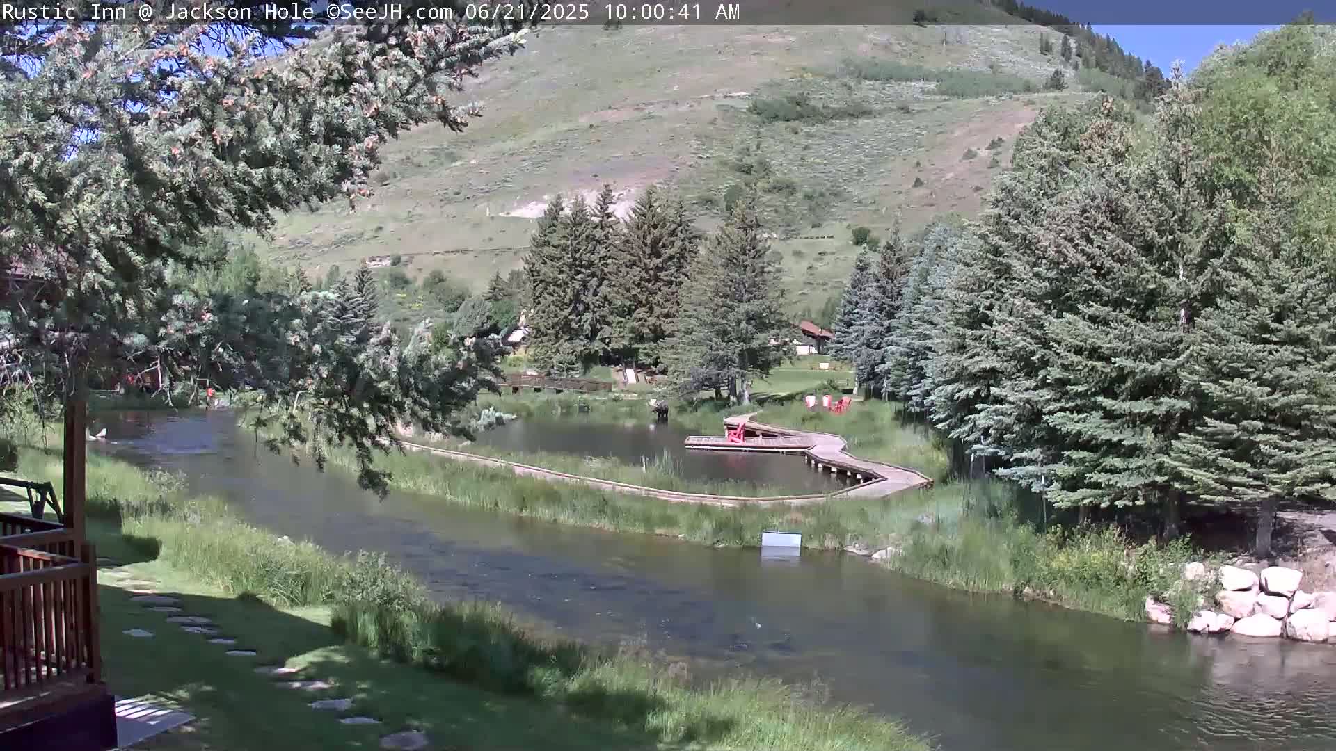 A calm river flows past a small pond with a wooden dock, surrounded by lush green vegetation and evergreen trees on a sunny day, with hills visible in the background.