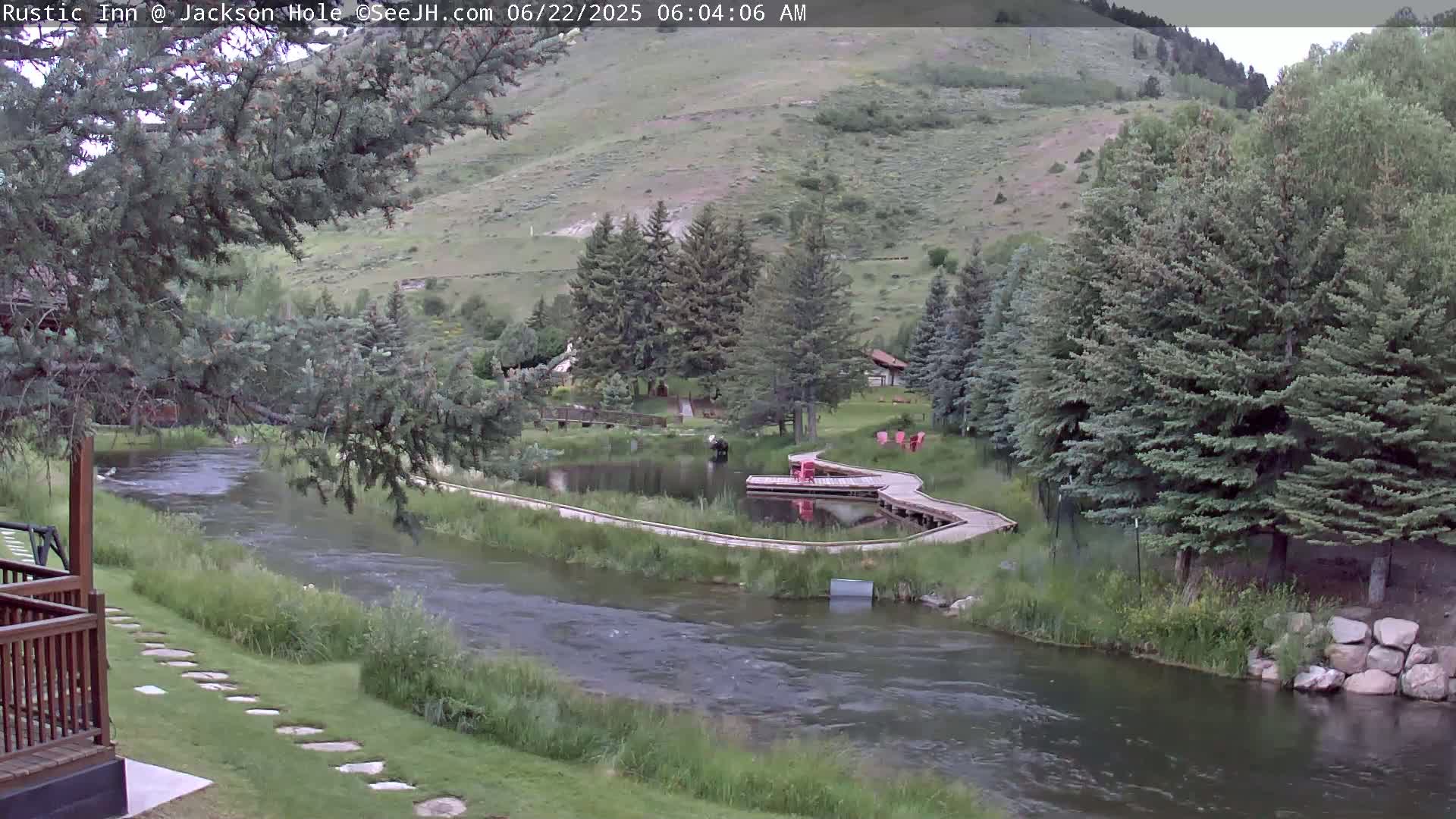A river flows through a green valley alongside a wooden walkway and small pond, surrounded by evergreen trees on a partly cloudy day.