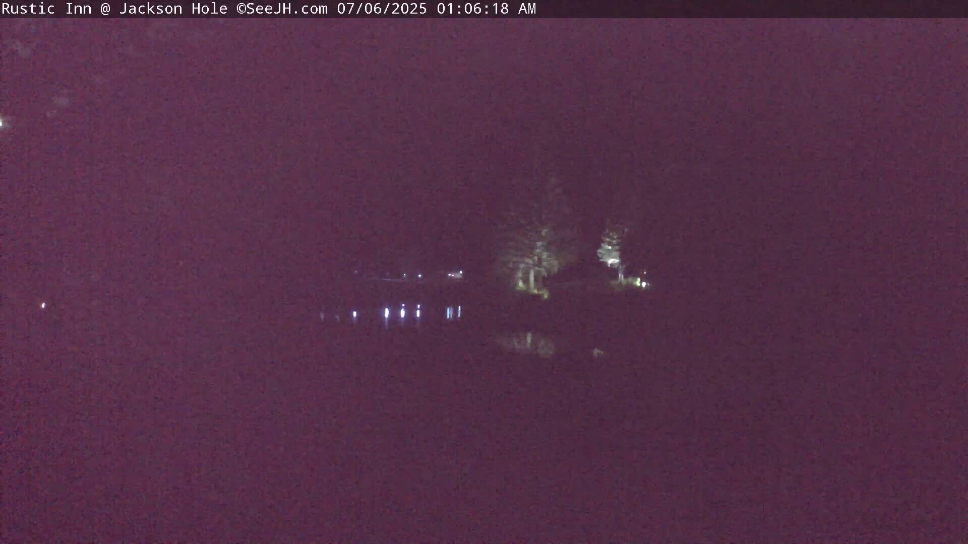 A dark, nighttime scene shows a body of water reflecting a few faint lights on the far shore, with some trees silhouetted against the dark sky.