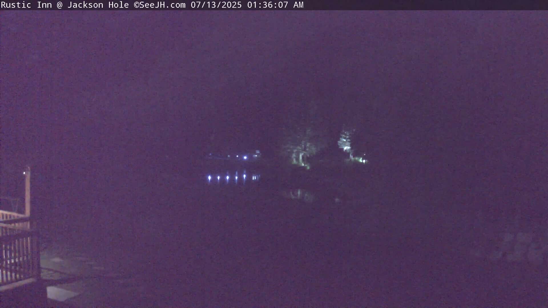 A dark nighttime scene shows a body of water reflecting some lights on the far shore, with trees and a portion of a wooden deck visible in the foreground.