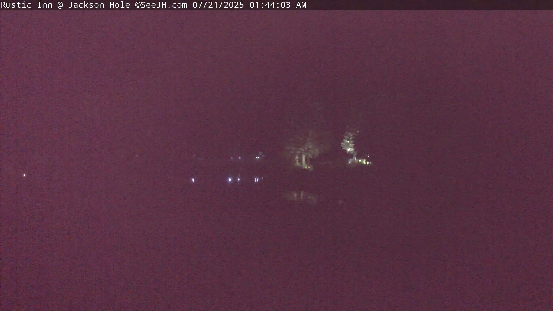 A nighttime, low-light image shows a dark body of water with a few lights visible on the far shore and some trees silhouetted against the dark sky.