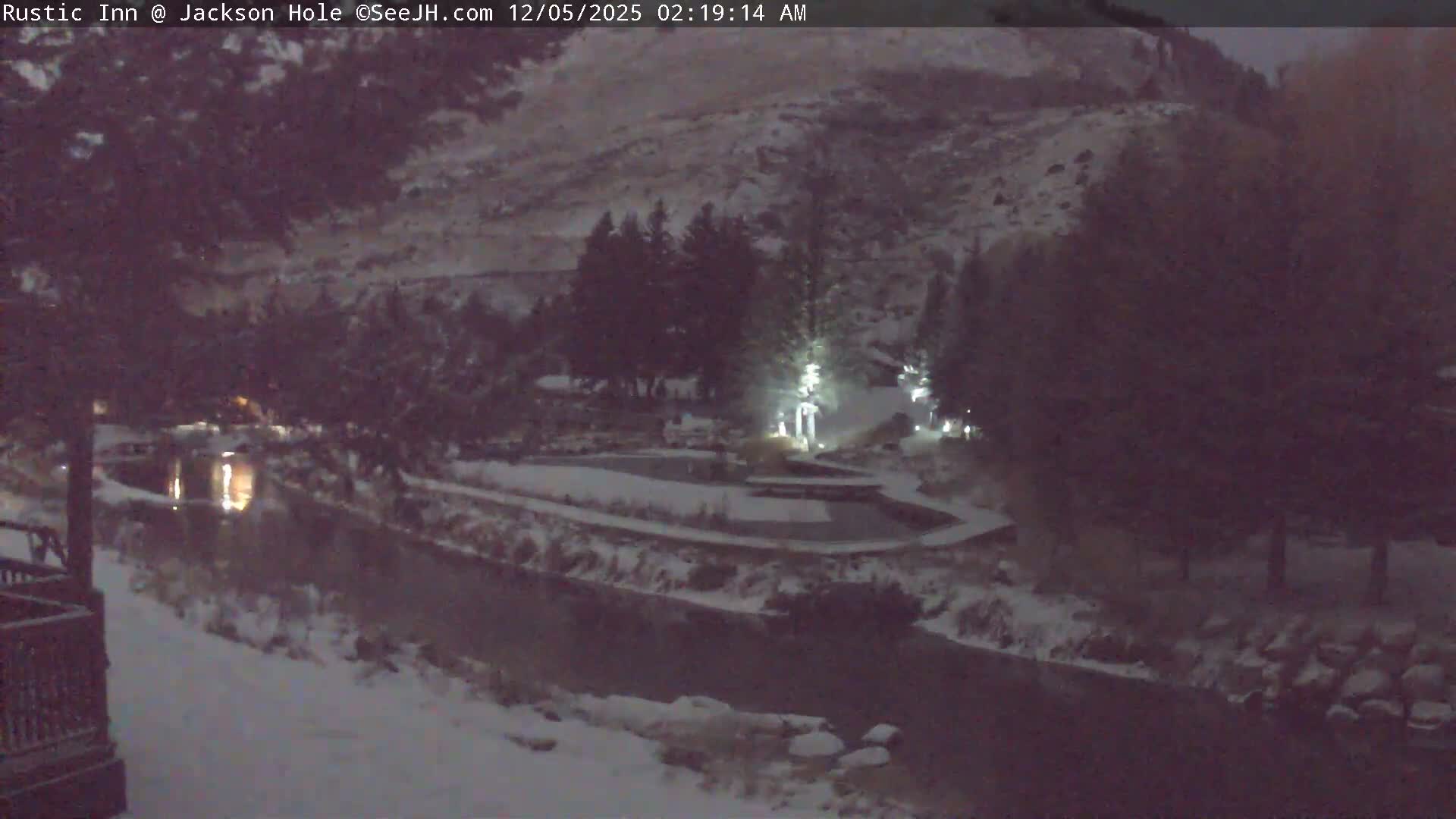 A clear, cold, snowy night scene shows a river reflecting warm lights, flowing past illuminated buildings and snow-covered trees, with snow-dusted mountains rising in the background.