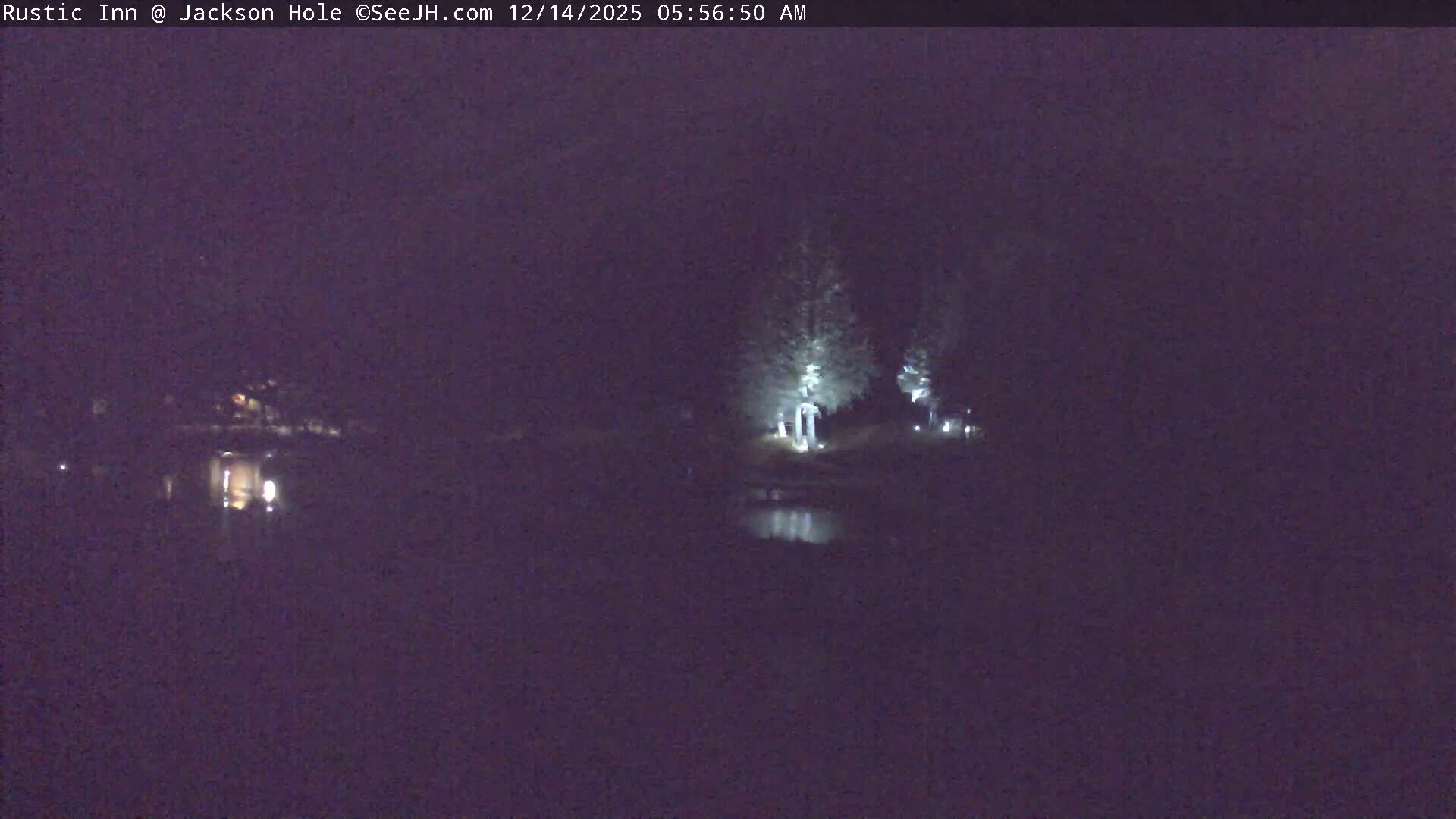 A clear, cold, snowy night scene shows a river reflecting warm lights, flowing past illuminated buildings and snow-covered trees, with snow-dusted mountains rising in the background.