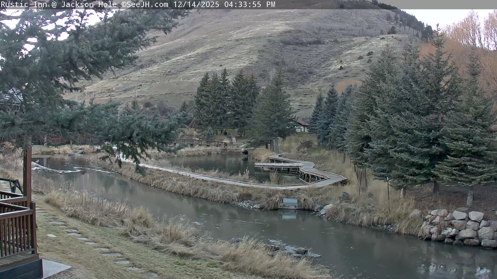 A clear, cold, snowy night scene shows a river reflecting warm lights, flowing past illuminated buildings and snow-covered trees, with snow-dusted mountains rising in the background.