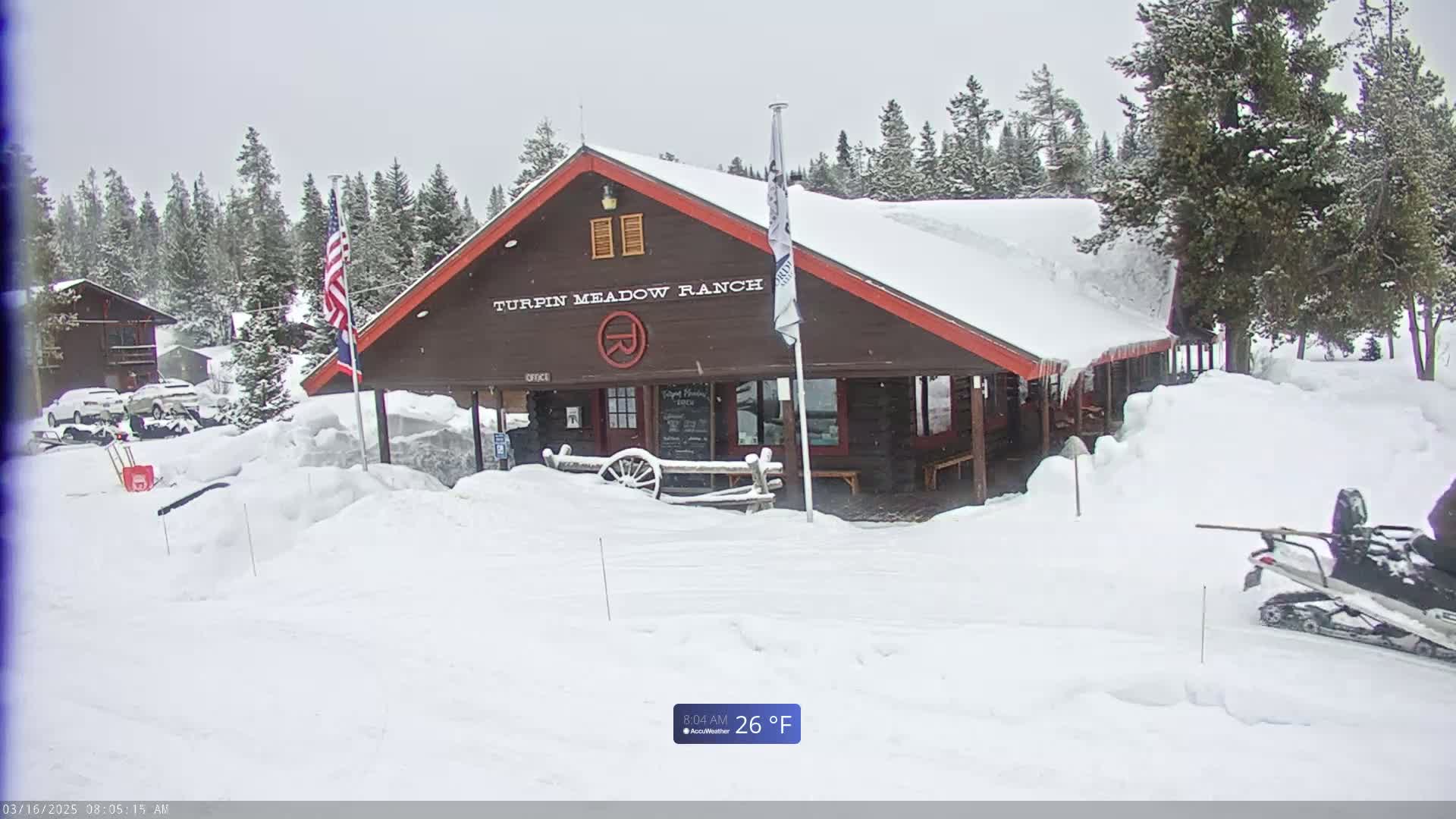 A snow-covered brown ranch building with red trim sits in a snowy landscape of evergreen trees, a snowmobile is parked near the building, and the temperature is 26°F.